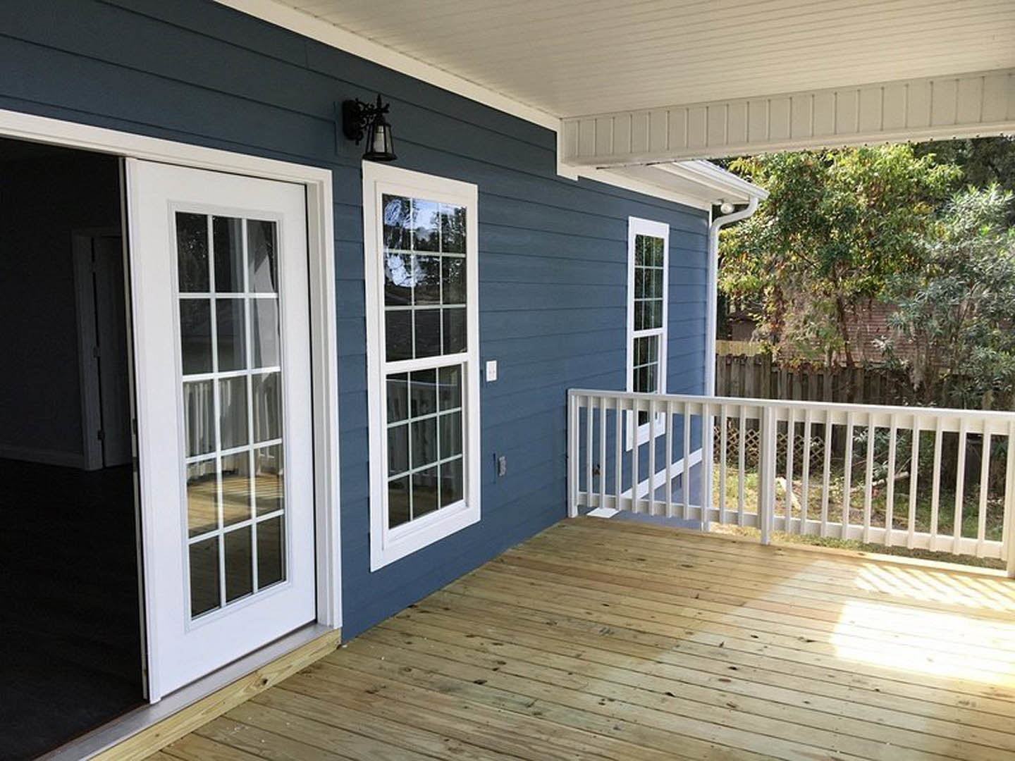 Wooden deck with white railing attached to a blue house, featuring a window and door, with a tree and fence nearby