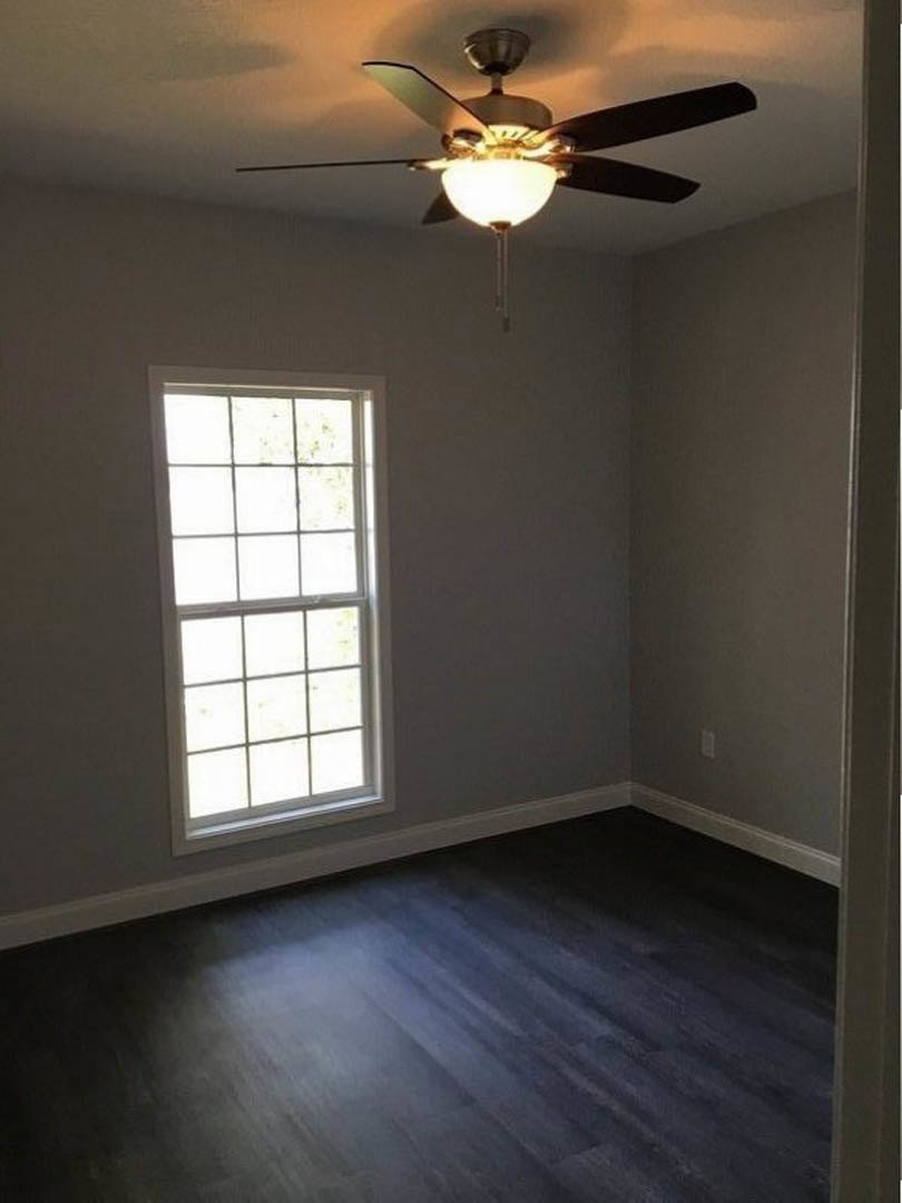 Ceiling fan with light fixture mounted on white plaster ceiling, dark wood flooring, large window with white trim, neutral walls