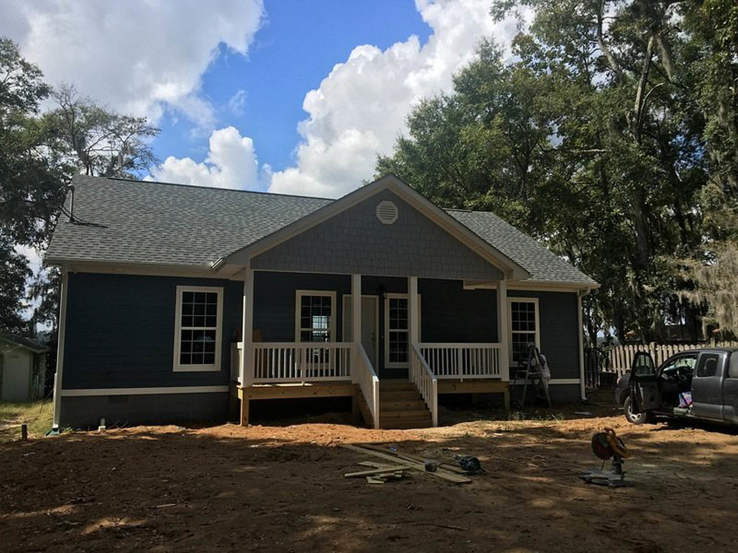 Two-story home with white siding, covered front porch, and stairs leading to a landscaped yard with mature trees; parked car with open door visible in driveway; Little White House