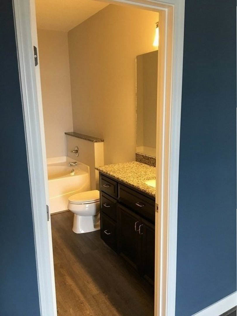White porcelain toilet and rectangular sink set against light gray tile walls, marbled countertop with dark wood cabinets, chrome faucet, and neutral-toned floor tiles.