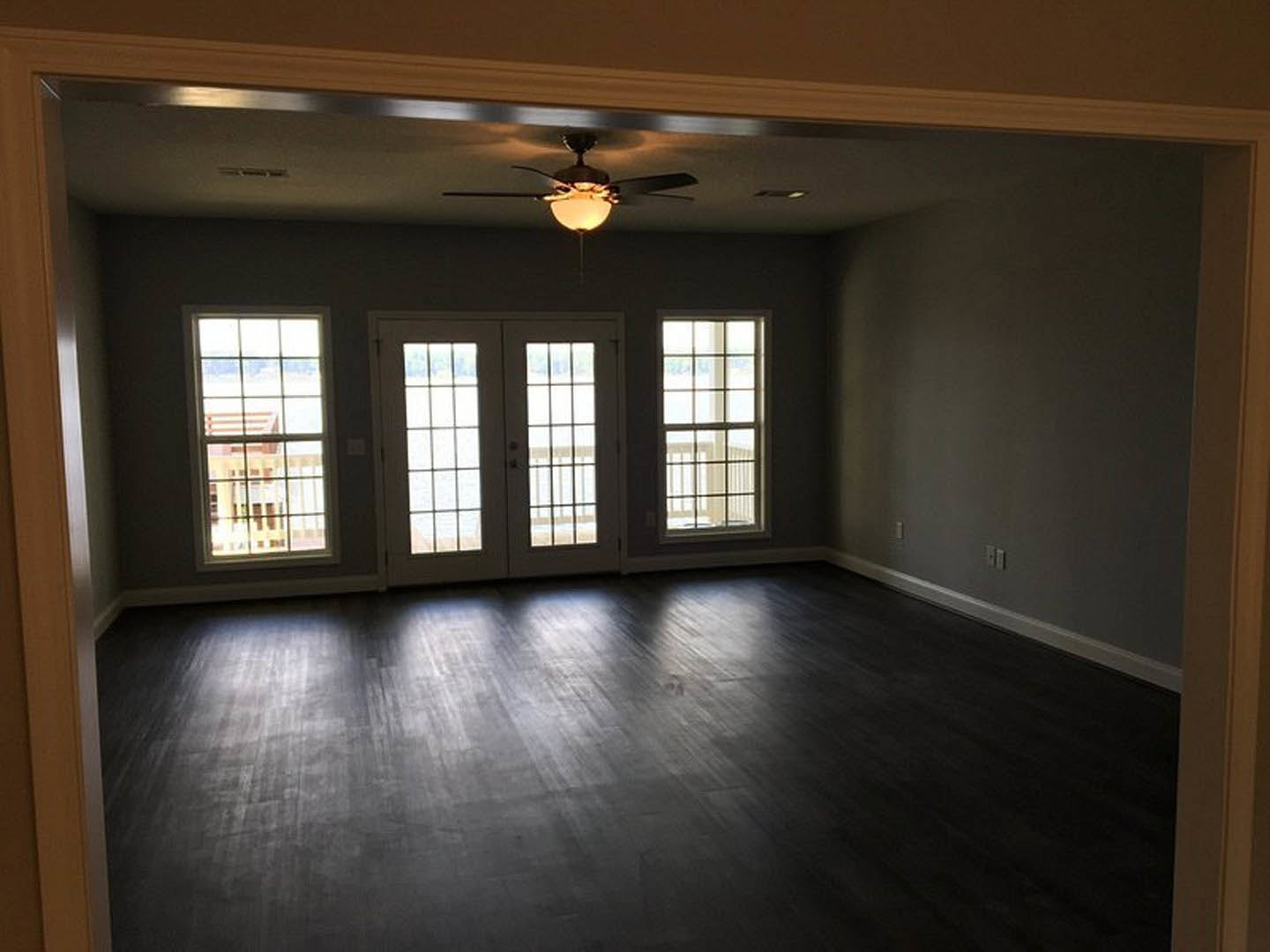 Dark hardwood floor reflecting sunlight, ceiling fan with illuminated light fixture, double glass-paneled doors, multi-pane windows including one with a railing, neutral walls and
