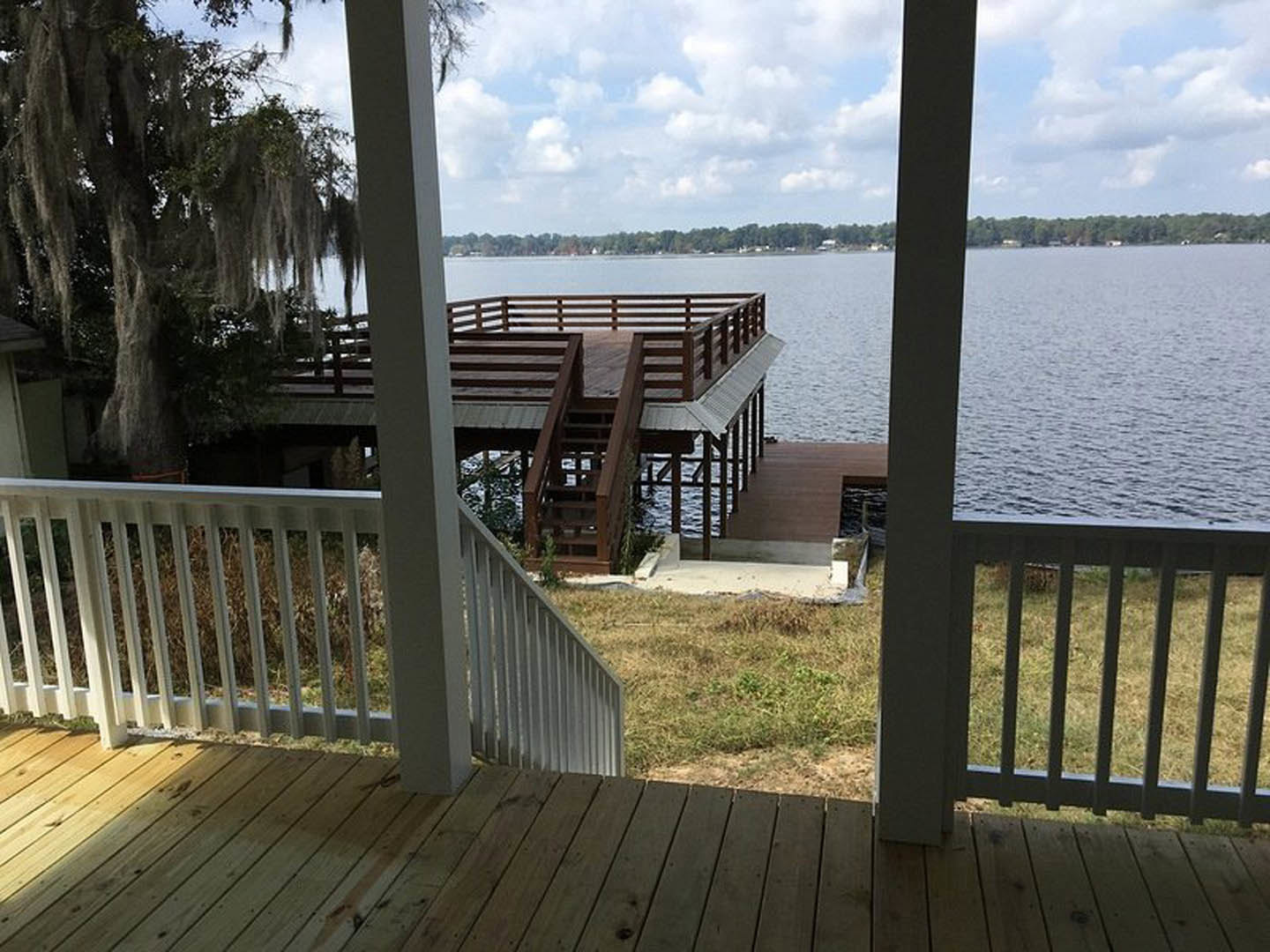 Wooden deck with white railing overlooking a lake, connected to a dock with metal railing; moss-covered tree and grassy area beside corrugated metal wall.