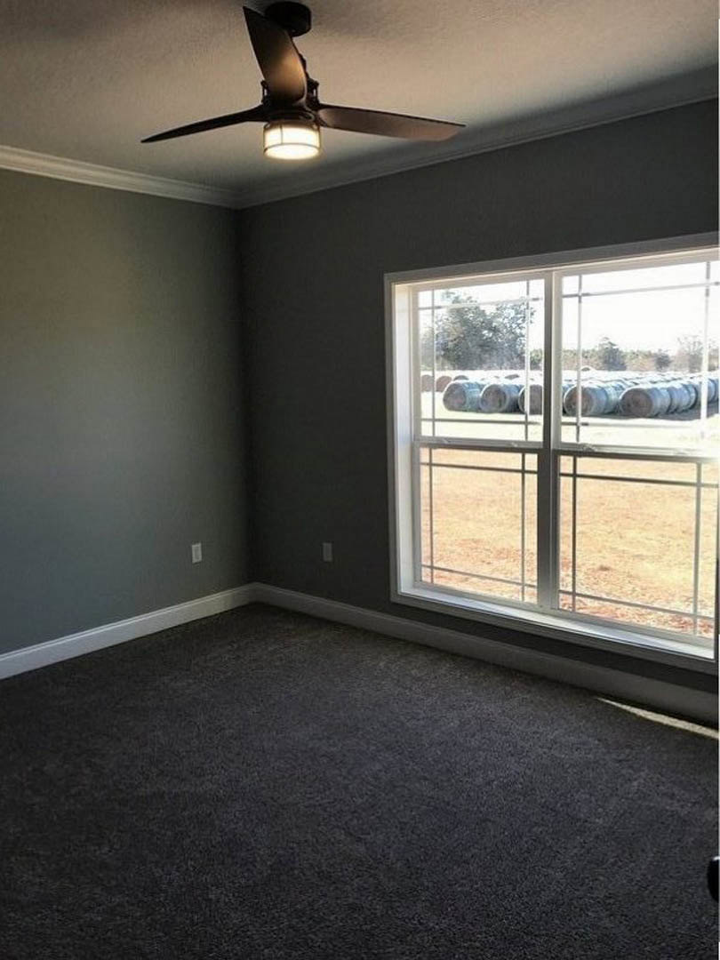 Carpeted room with white walls, ceiling fan and light fixture, large window overlooking field of barrels