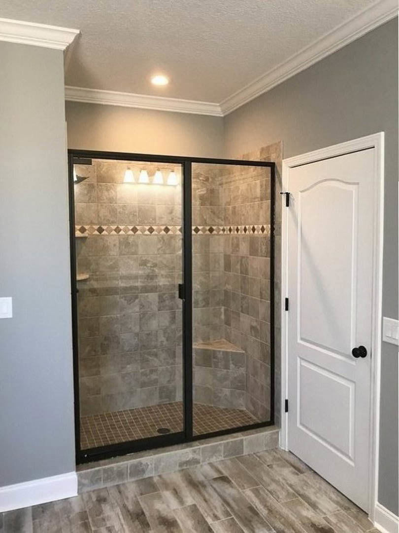 Shower with clear glass door and black frame, white paneled door with black knobs, grey tile floor bordered in black, white walls with black trim