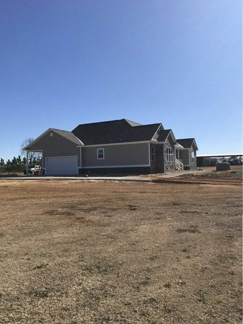 Two-story house with black roof, attached garage with open door, surrounded by large dirt field and patches of brown grass under clear blue sky