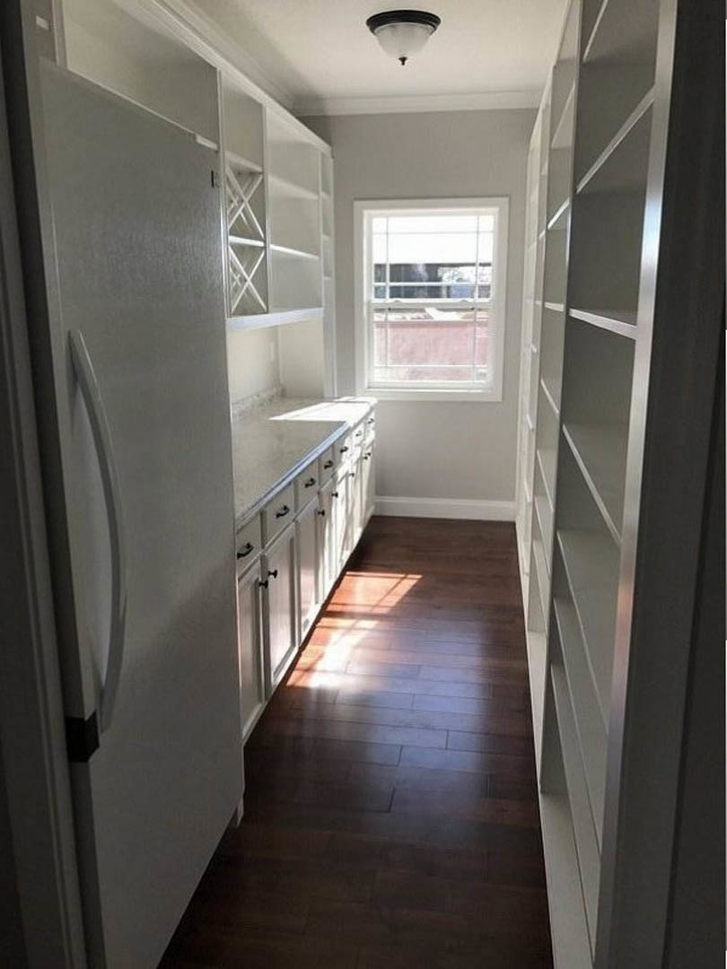 White cabinetry and open shelves in a kitchen with dark wood flooring, stainless steel refrigerator, pendant light fixture, and a window overlooking a neighboring building.