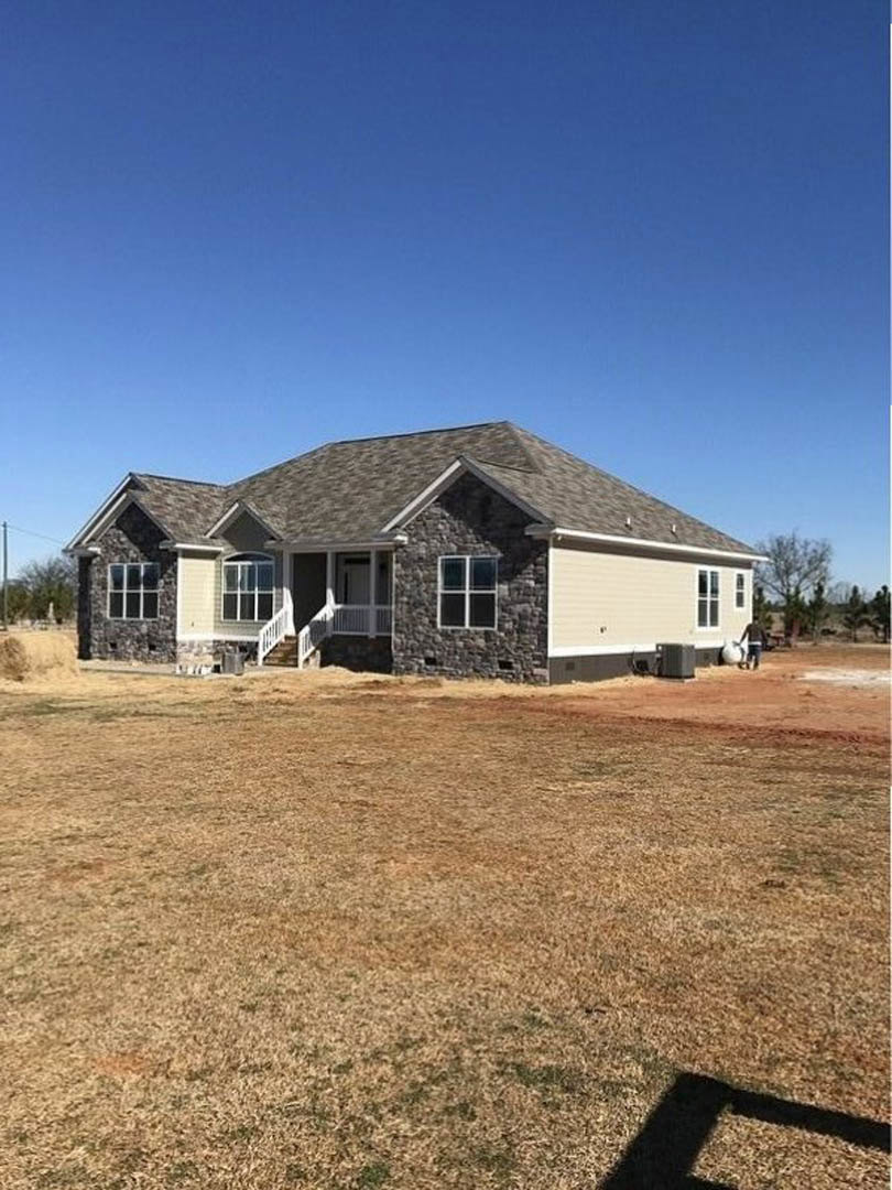 Two-story house with stone exterior wall, white-framed windows, and expansive grassy yard under clear blue sky; shadow of a person visible on lawn.