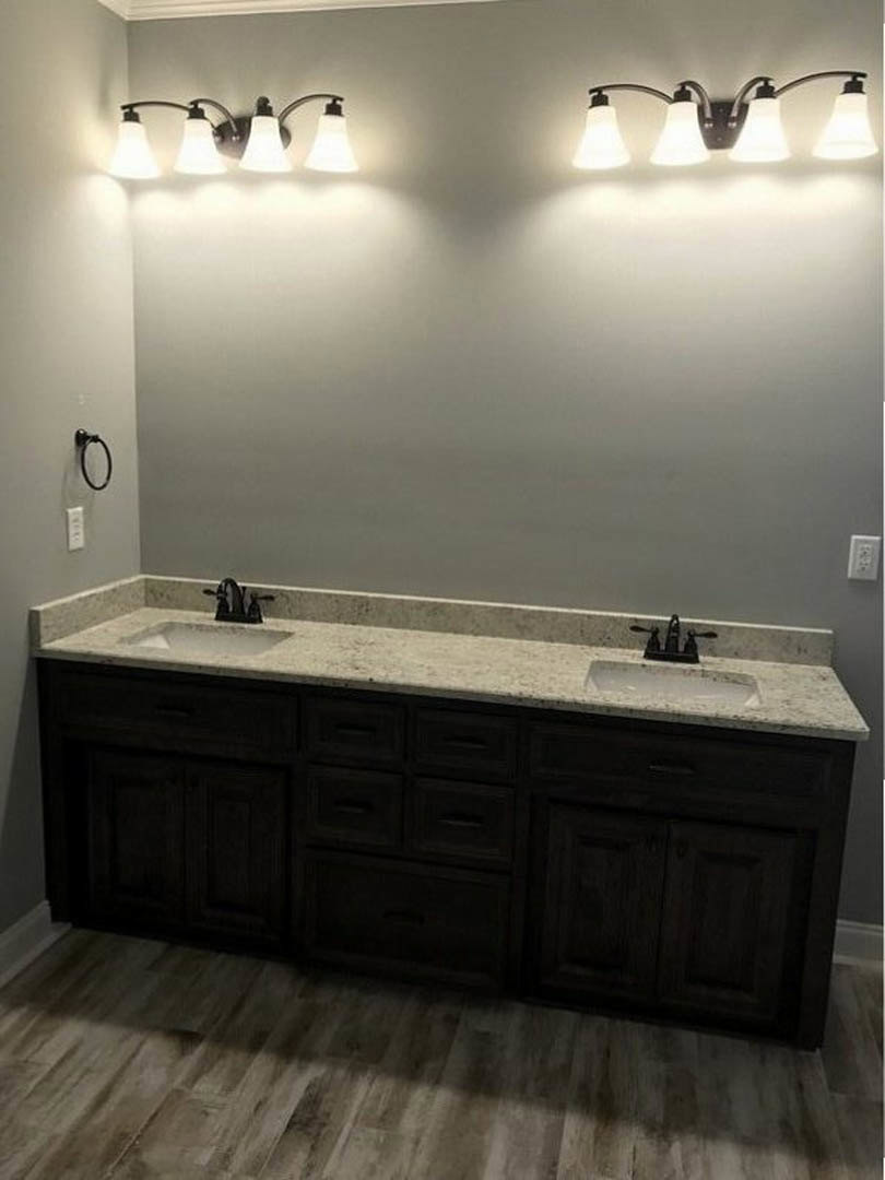 Double vanity with white countertop and dark wood drawers, wood plank flooring, two modern light fixtures above sinks, gray wall backdrop