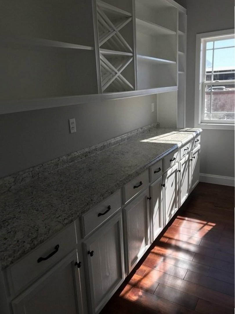White kitchen cabinets with black handles, open shelves, wood flooring, tile backsplash, and a window overlooking a parked car.