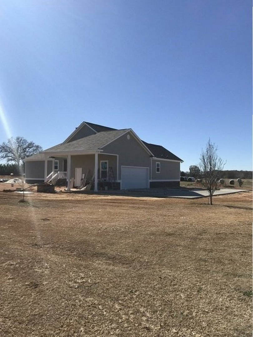 Partially built house with exposed framing, attached garage, white door and window frames, leafless tree nearby, situated in an open grassy field under clear blue sky