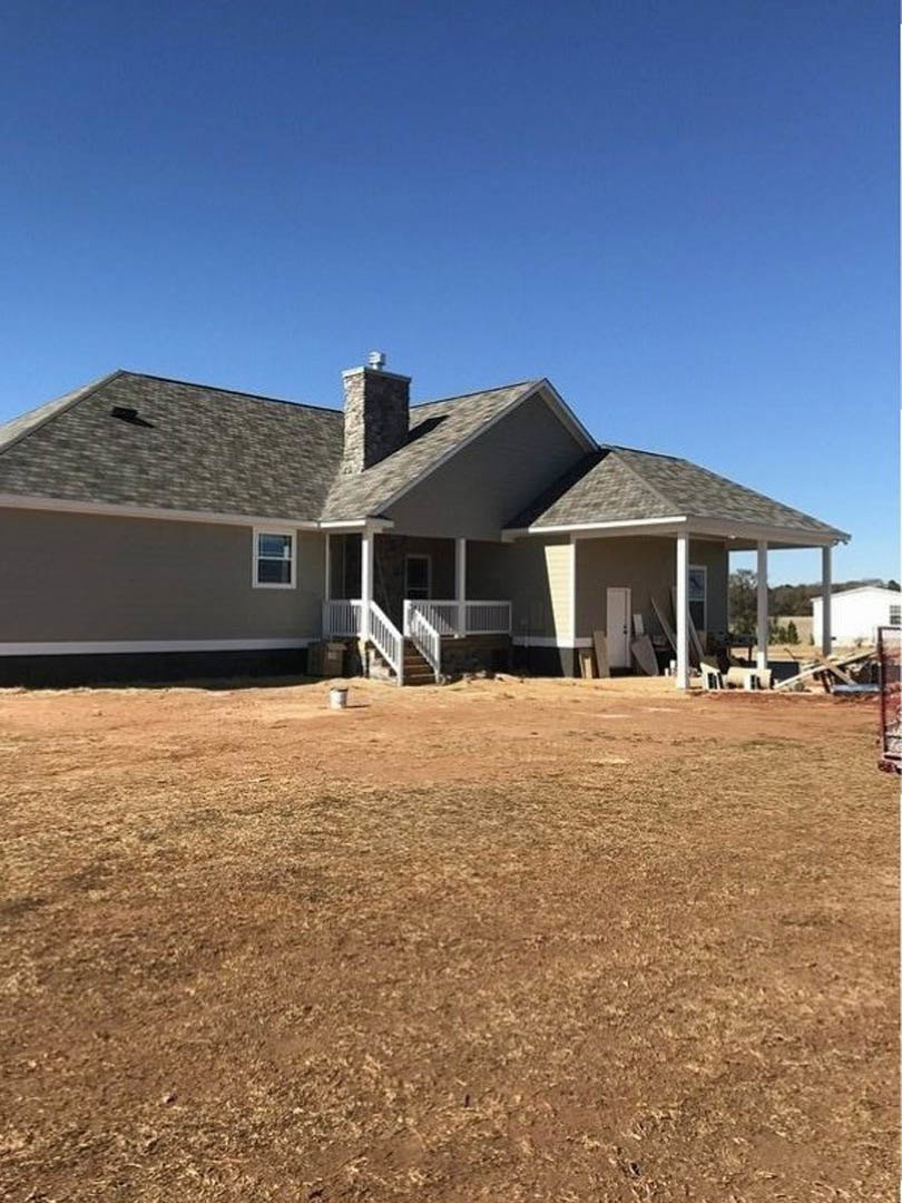 White house under construction with exposed chimney, brown dirt field in foreground, white porch railing, and unfinished exterior walls.