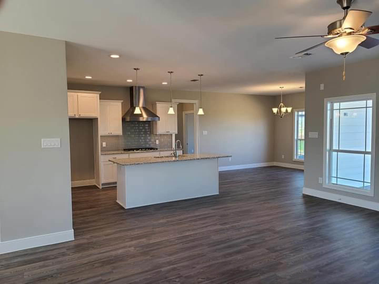 Open-concept kitchen and living room featuring hardwood floors, white walls, large window with white frame, kitchen island with cabinetry, stainless steel vent hood, and recessed