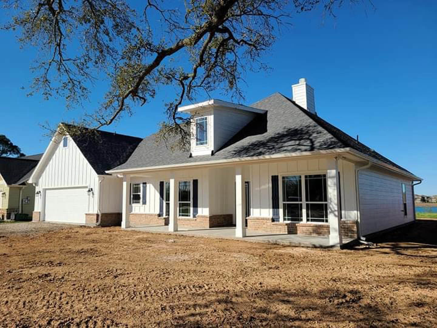 White house with black roof and blue-trimmed windows, tree and blue sky in background, dirt field in foreground