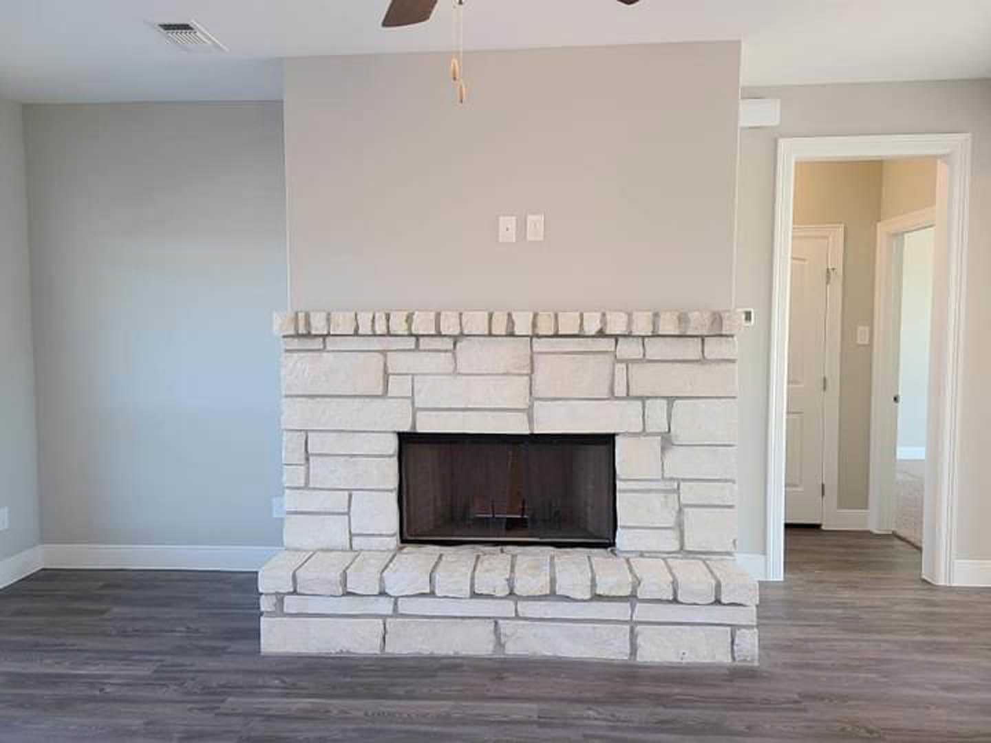 Stone fireplace set in a brick accent wall, wood-burning stove with fire screen, hardwood floor, white walls, hallway with white doors, window nook featuring a chair and sheer