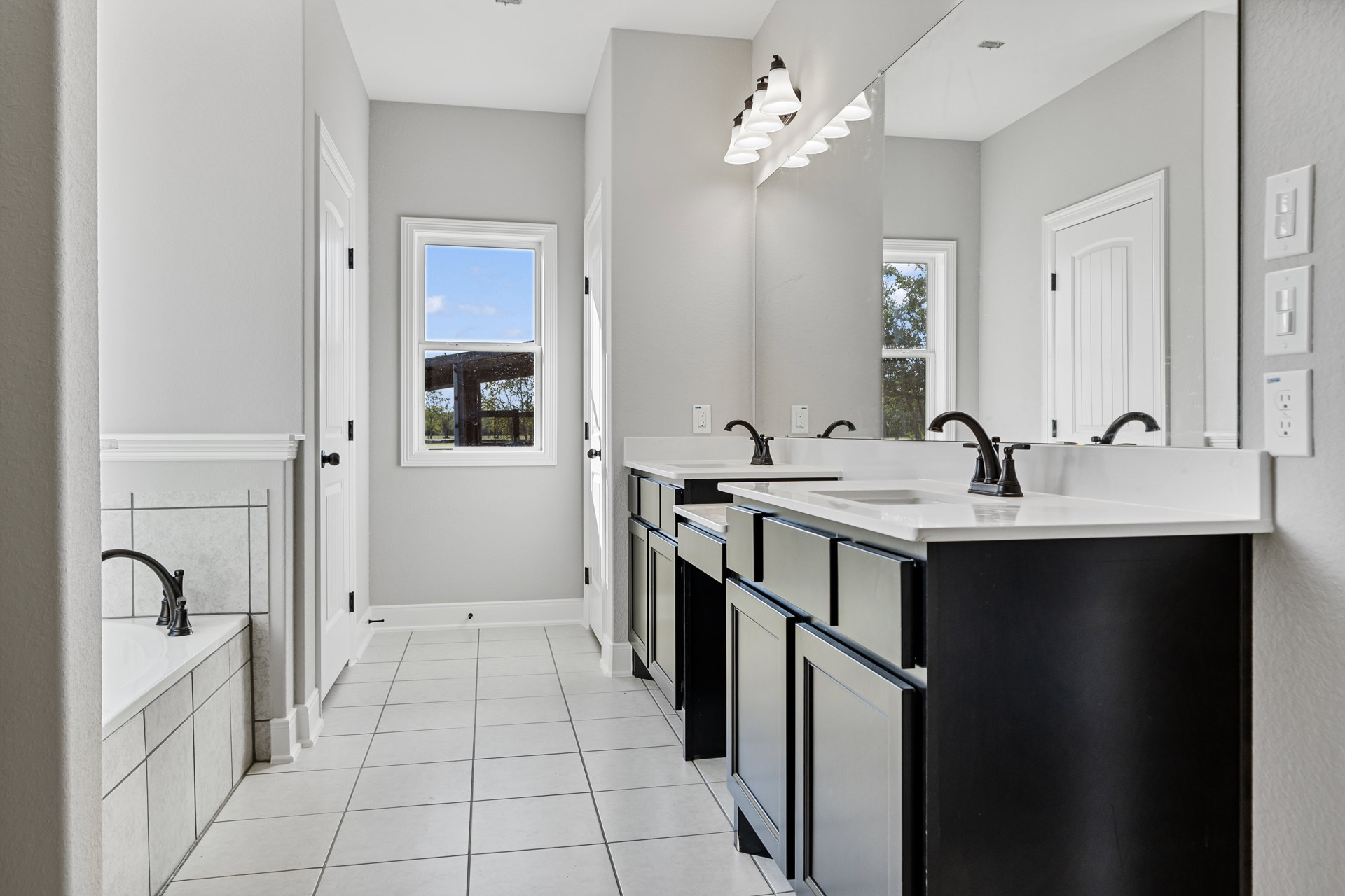 Bathroom with expansive mirror above dual sinks, black faucets on white countertops, white cabinetry, white tile floor, window showing blue sky, white door with black hardware
