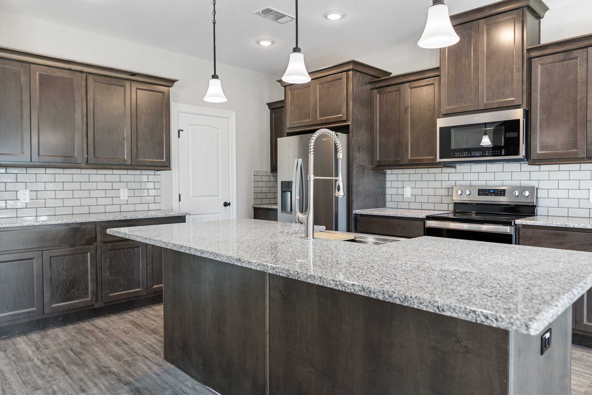 Granite island with white cabinetry, stainless steel microwave and oven, undermount sink with silver faucet, pendant light fixture with white shade