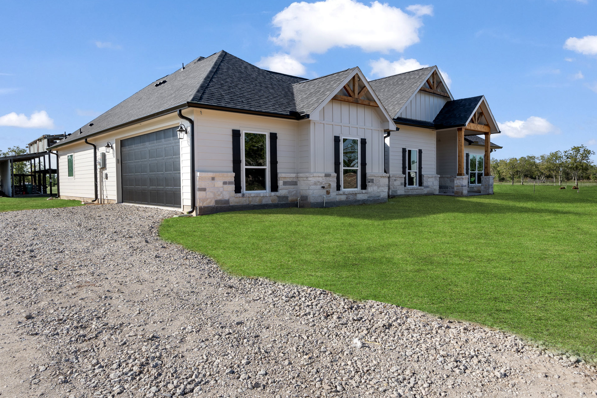 Gravel driveway bordered by green lawn leading to a house with black roof, white-framed windows, and garage door topped by exterior light; partly cloudy sky overhead.