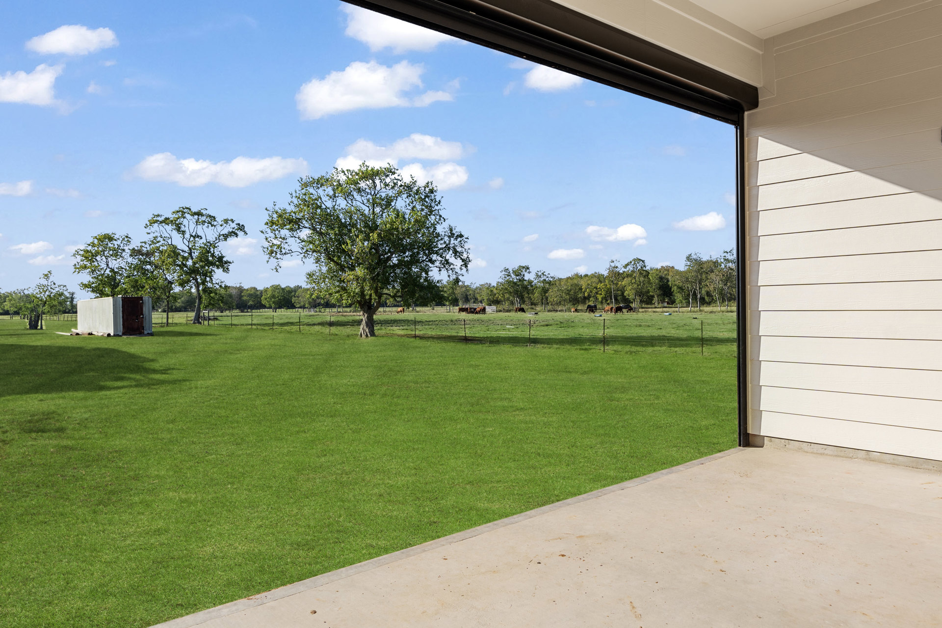Wide open brown door in white siding wall, concrete walkway leading to expansive green lawn with scattered trees and distant building under partly cloudy sky