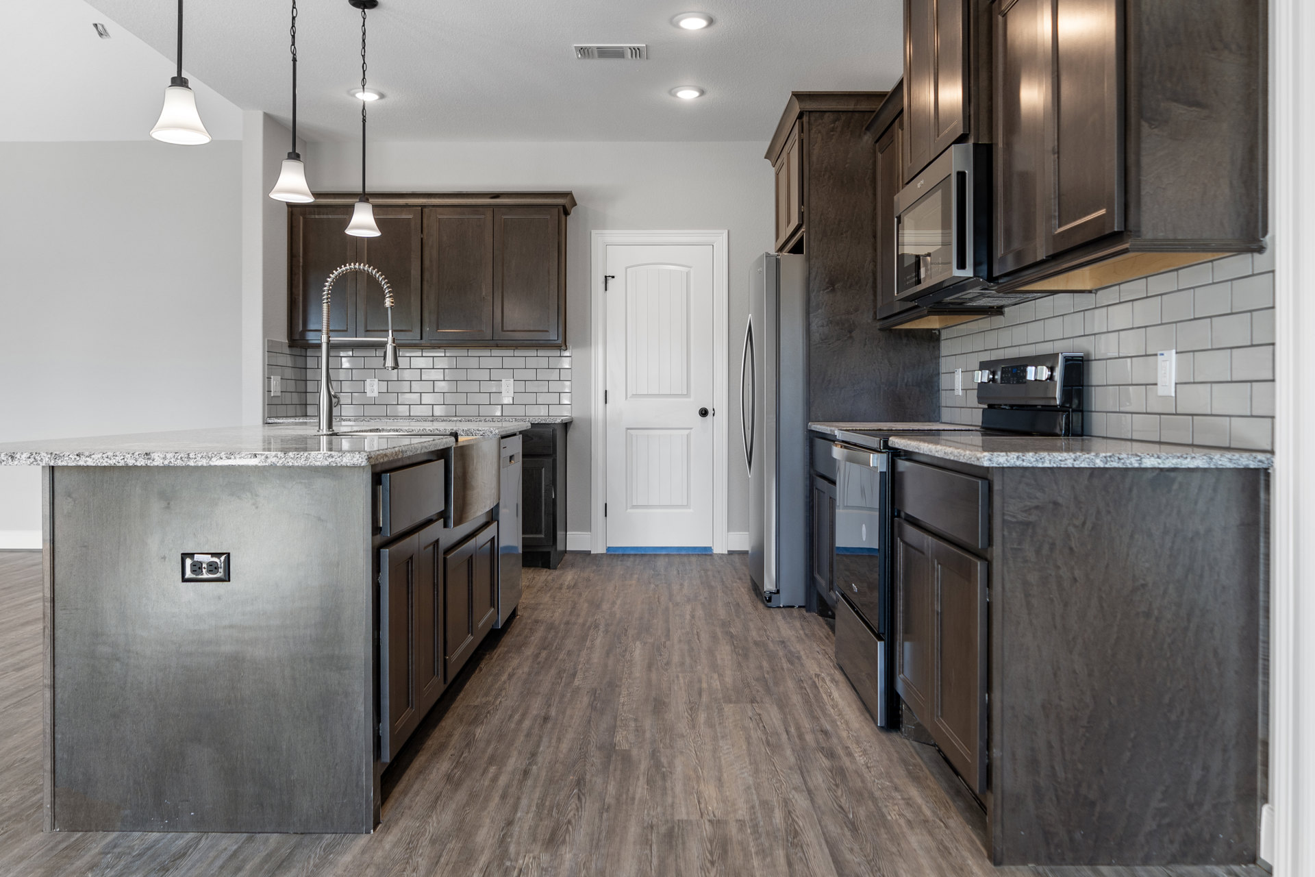 Kitchen with dark wood cabinets, wood flooring, white door with black knob, kitchen island featuring power outlet, stove close-up, and light fixture with white shade