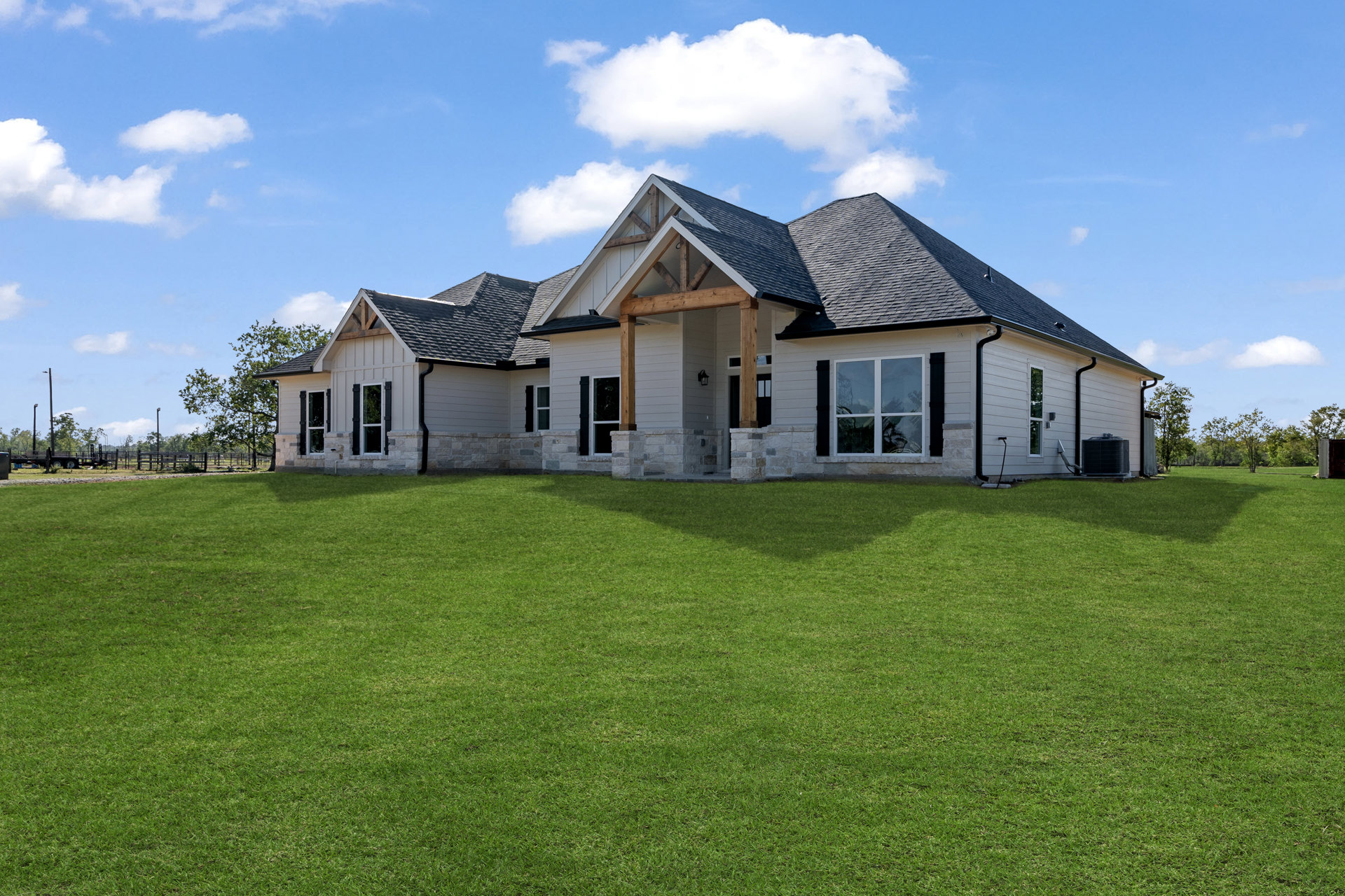 Expansive green lawn leading to a stone farmhouse with white-framed windows, wood beams, and a wooden porch beneath a partly cloudy sky