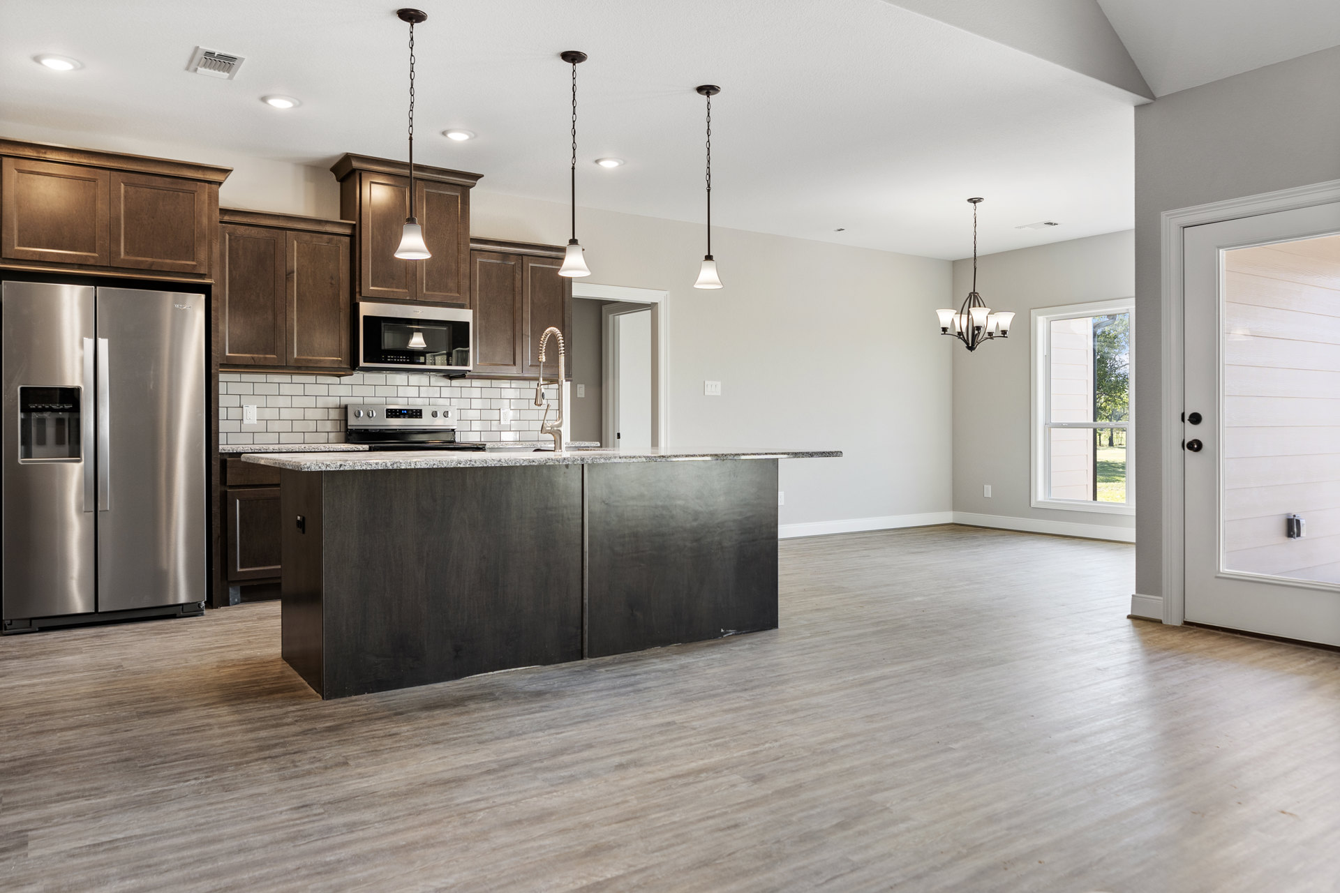 Open kitchen and living room featuring wood flooring, black marble countertop, stainless steel refrigerator, built-in microwave, white-shaded light fixture, and modern cabinetry.