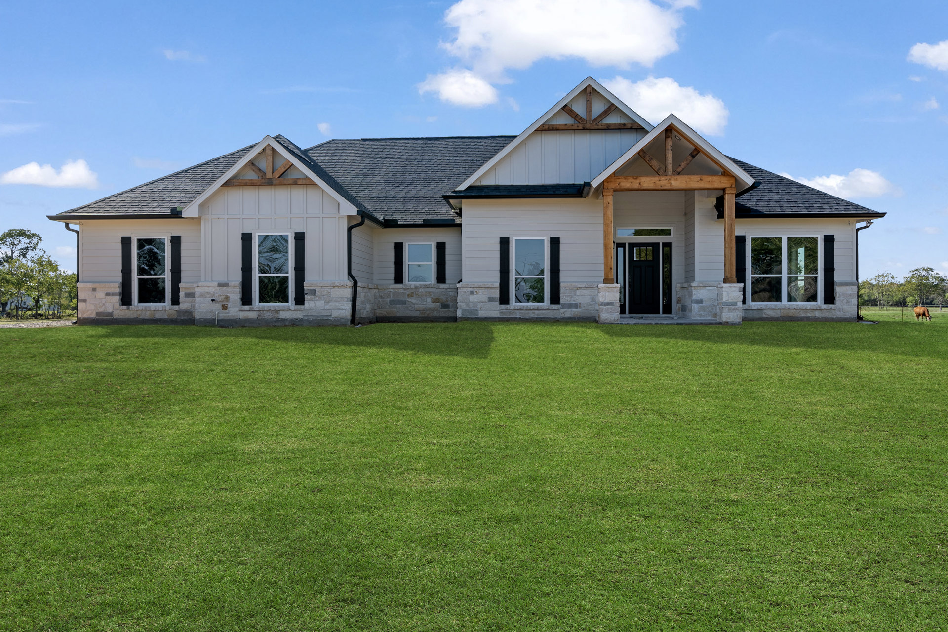 Two-story home with gray stone exterior, black front door, large windows, and manicured green lawn under a partly cloudy sky