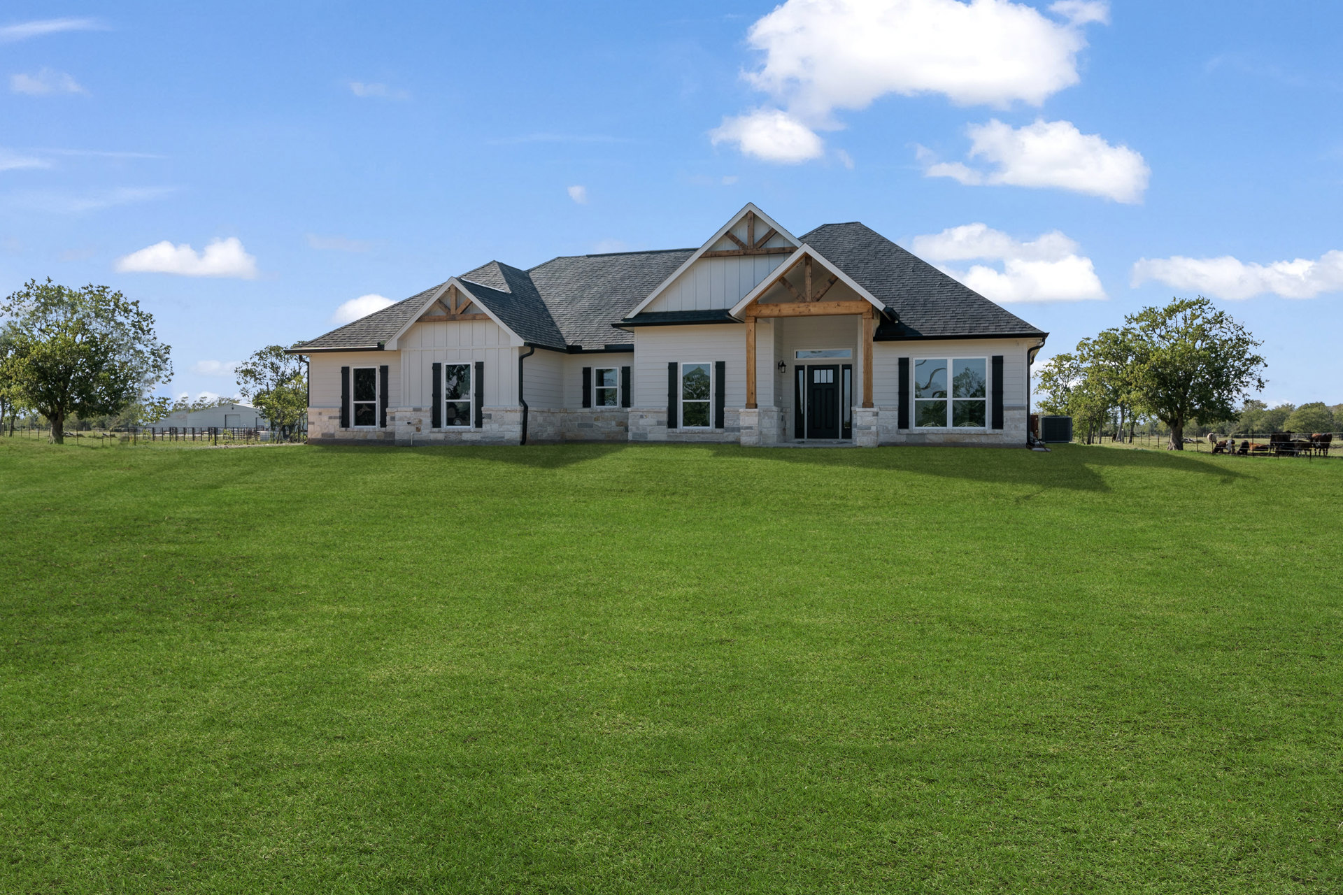 Two-story farmhouse with black front door and glass panes, wide covered porch, large green lawn, mature trees, and windows reflecting surrounding landscape