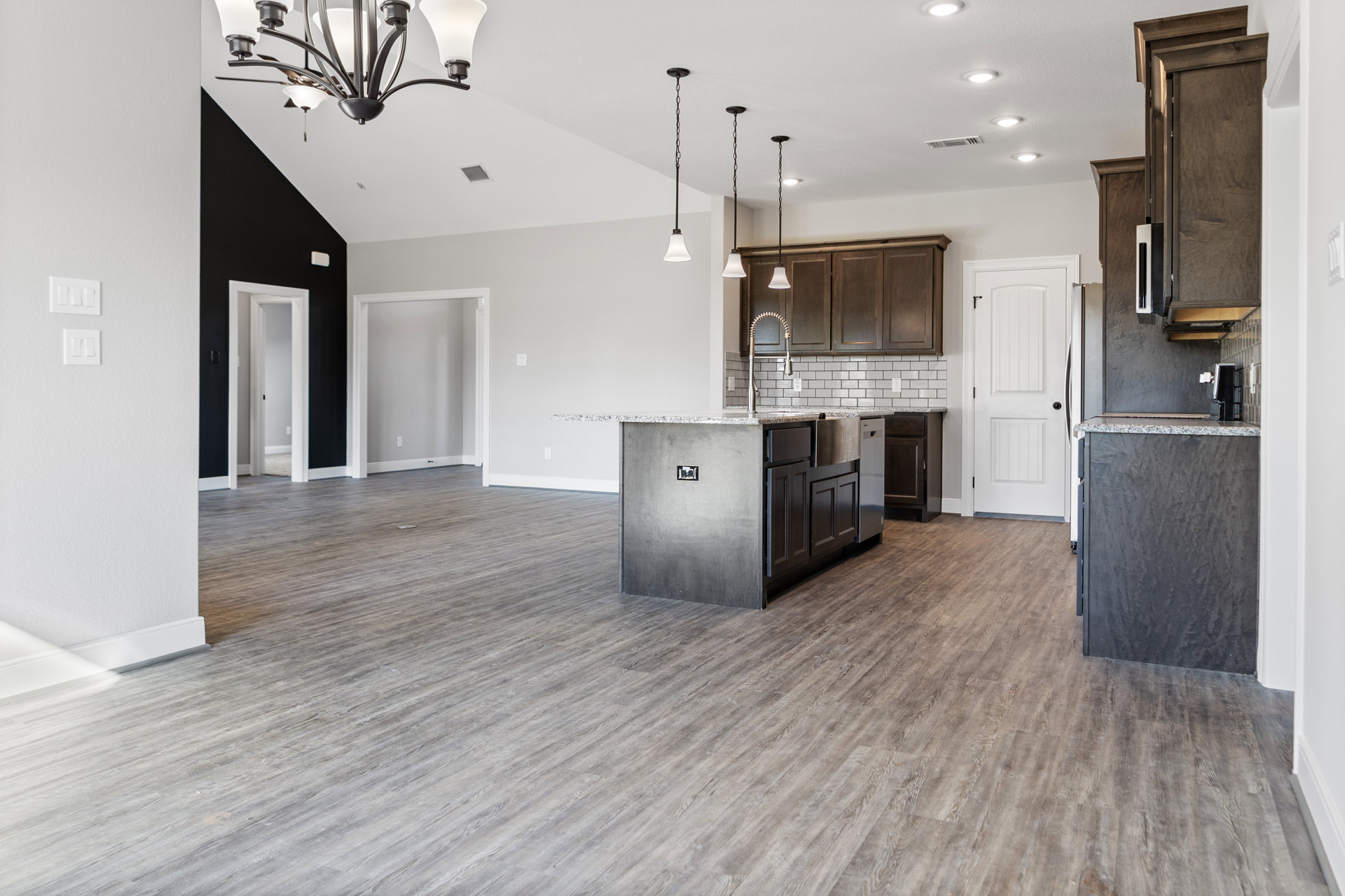 Open kitchen and living room featuring wood flooring, white cabinetry, kitchen island with built-in sink and faucet, black countertop, and modern chandelier above the island.