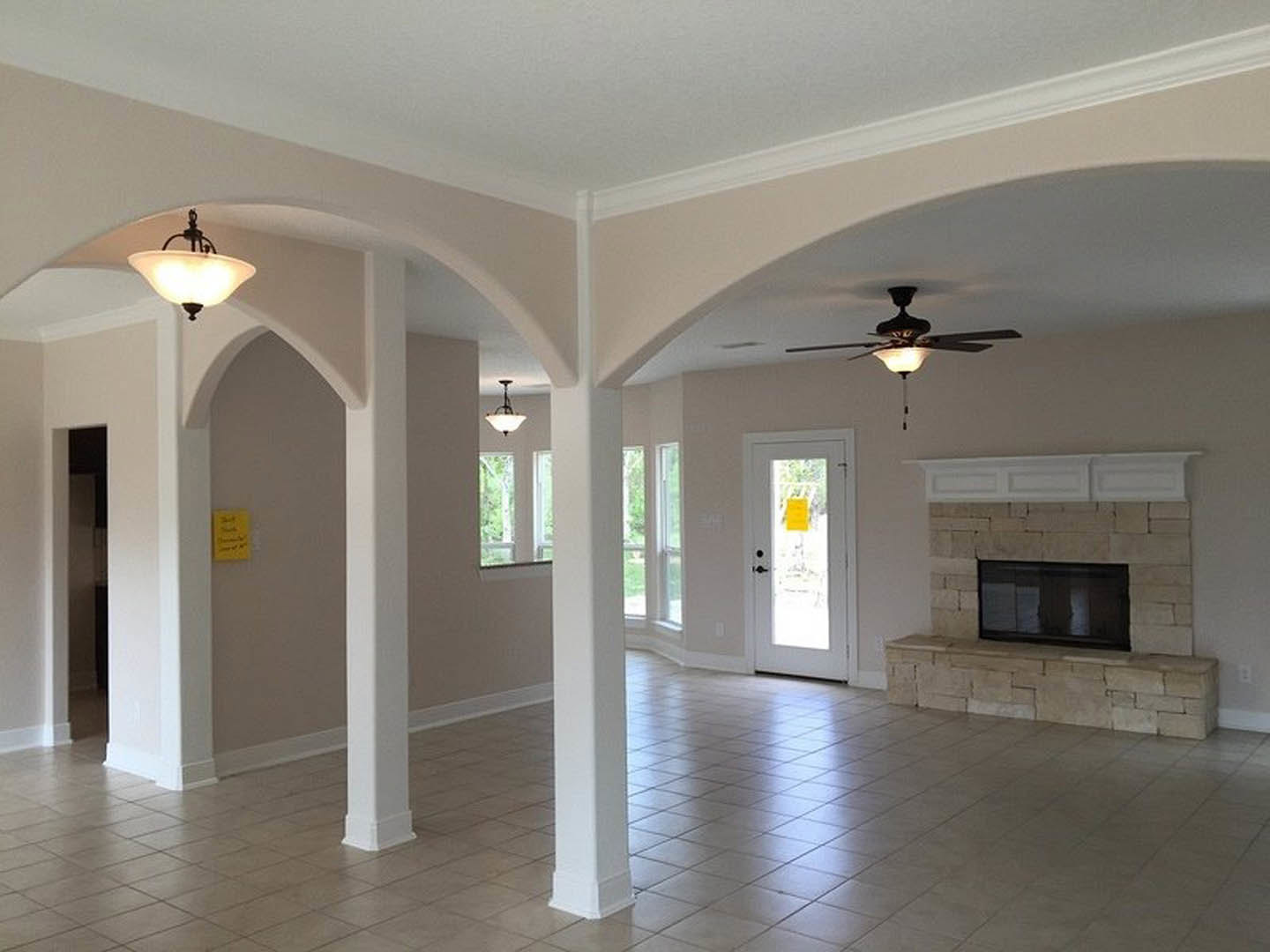 Spacious living room featuring a stone fireplace, ceiling fan with light, wood flooring, crown molding, and a door displaying a yellow sign with black writing; reflection of a