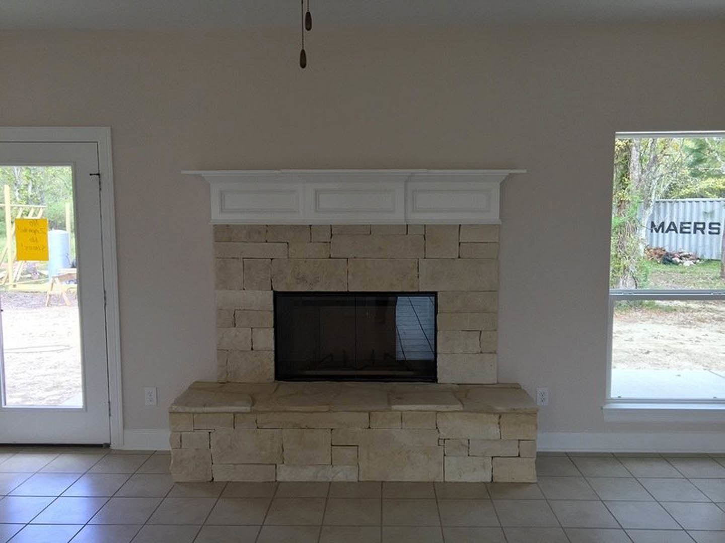 Stone fireplace with wood mantel, white walls with crown molding, large window reflecting a boat on the water, yellow sign posted on window, hardwood floor