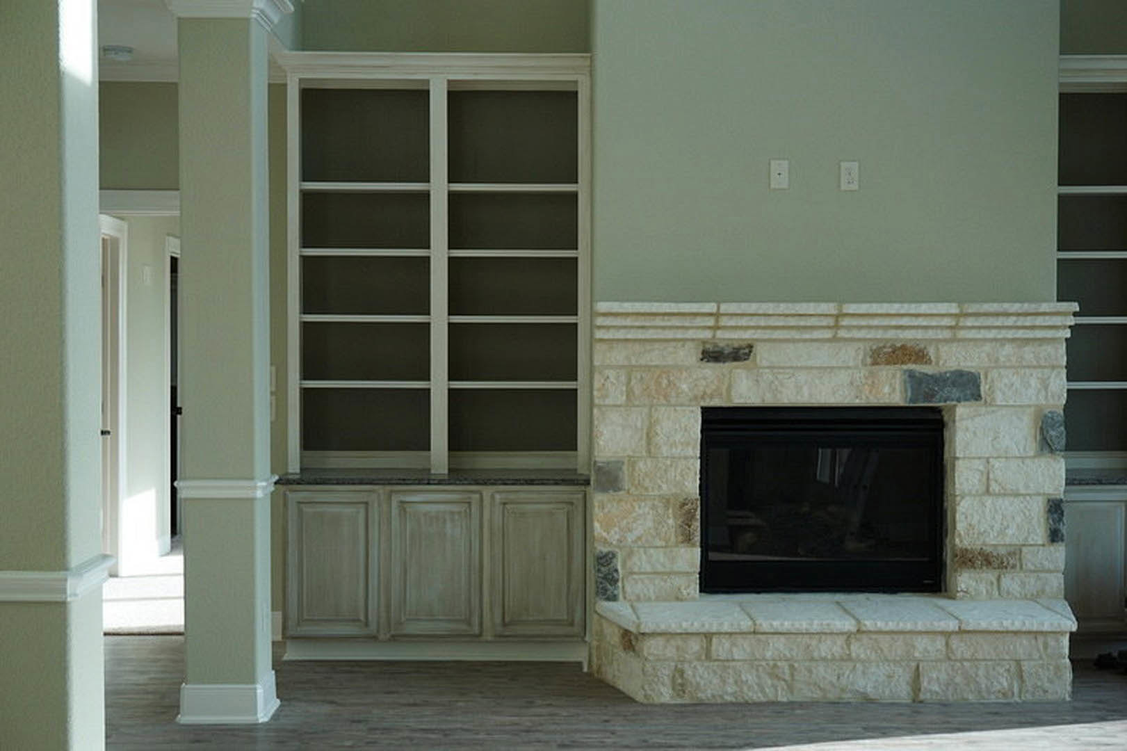 Stone fireplace with black glass front, white built-in bookcase with shelves, dark wood cabinet, large window, light hardwood floor, textured stone wall