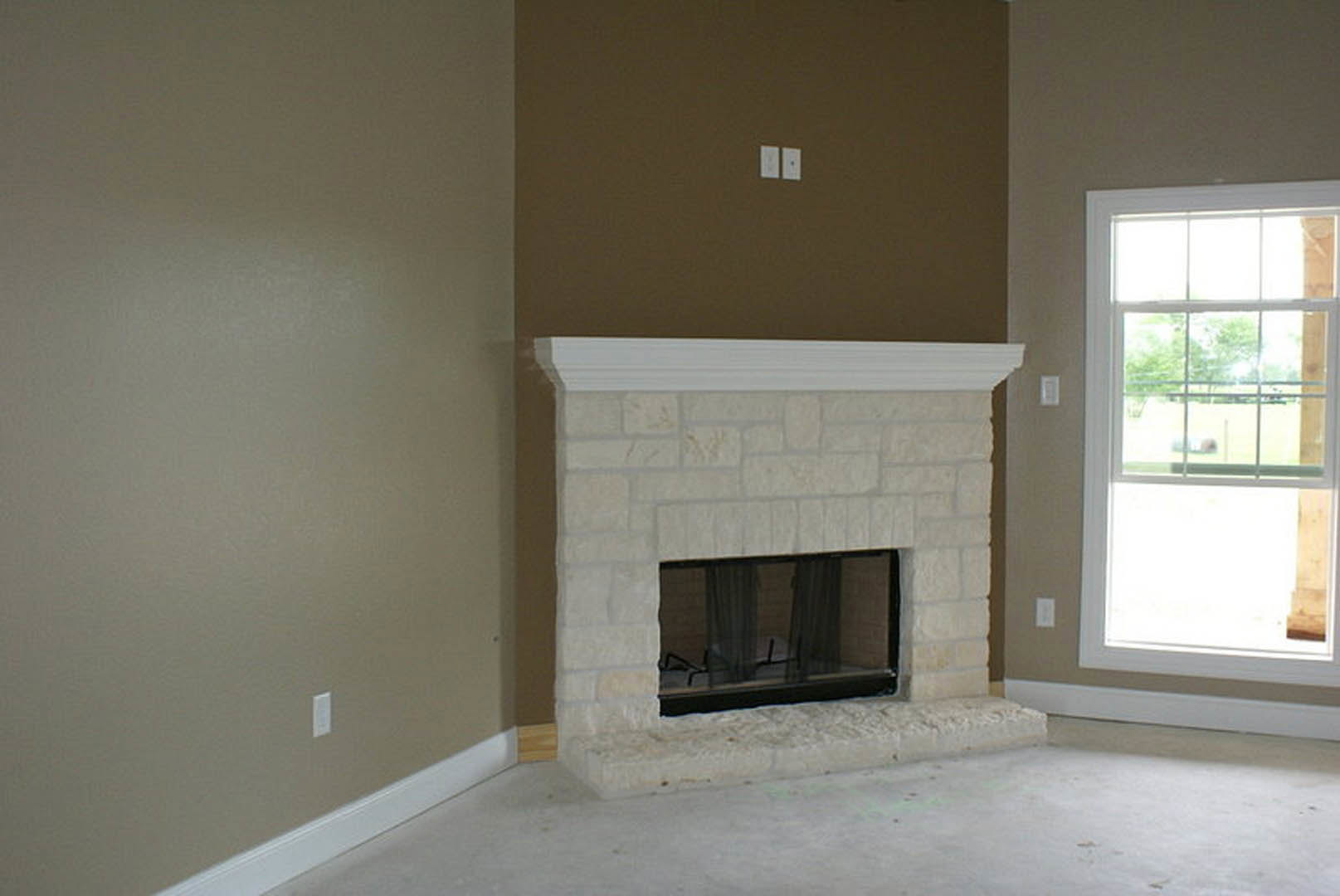 White stone fireplace with wood mantel, flanked by windows with sheer curtains, overlooking grassy field; plush white carpet covers floor.