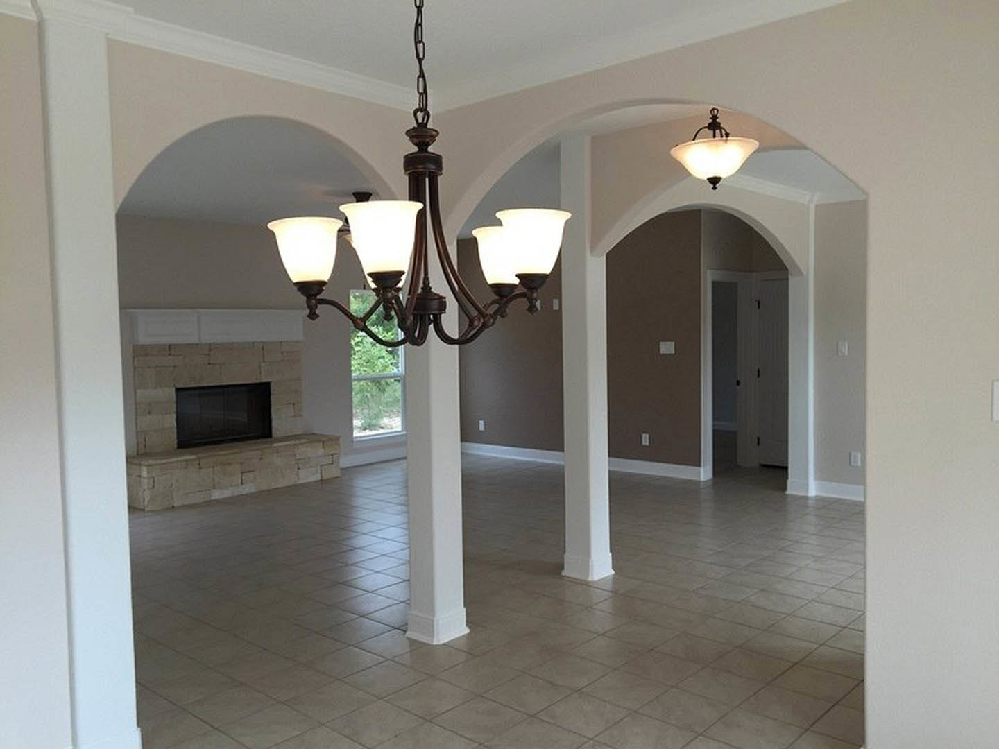 Elegant chandelier with multiple lights hanging from a coffered ceiling above a spacious living room featuring white pillars, tile flooring, large windows, and a stone fireplace.
