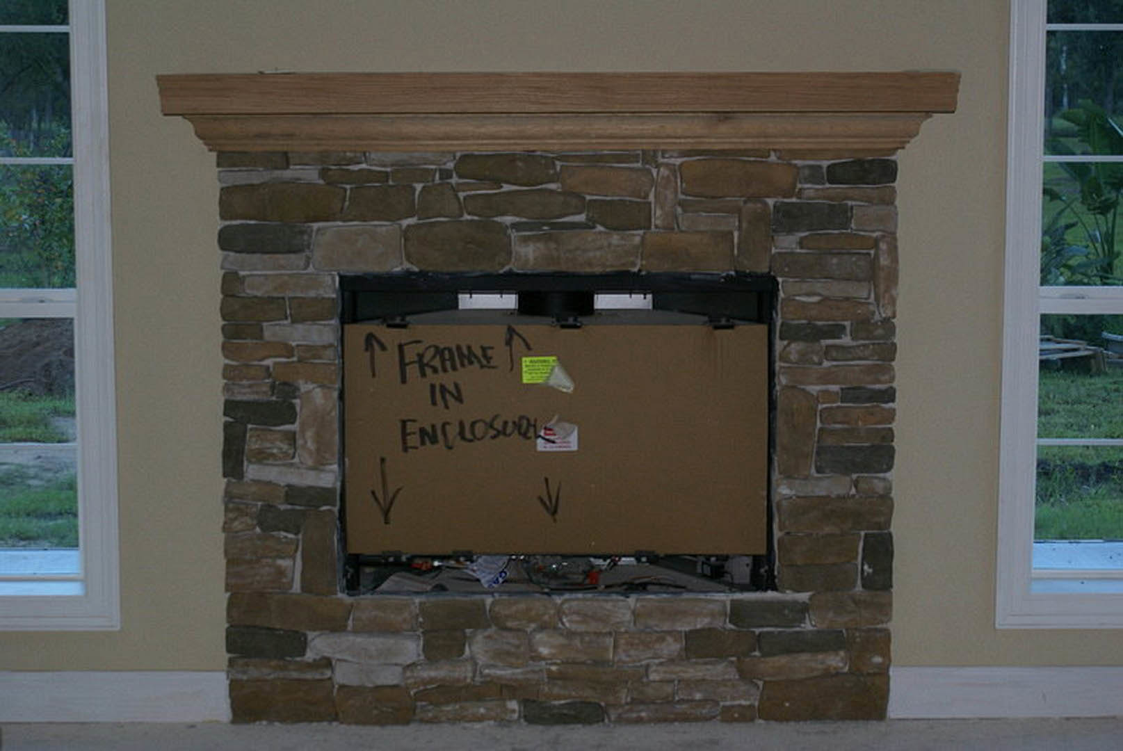 Brick fireplace with a cardboard box featuring black writing placed inside, surrounded by white walls and nearby windows