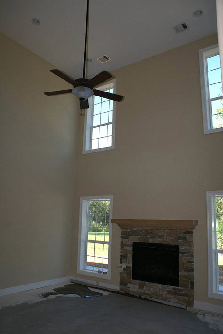 Ceiling fan with light fixture above living room featuring stone fireplace, large window overlooking grassy yard, and neutral walls