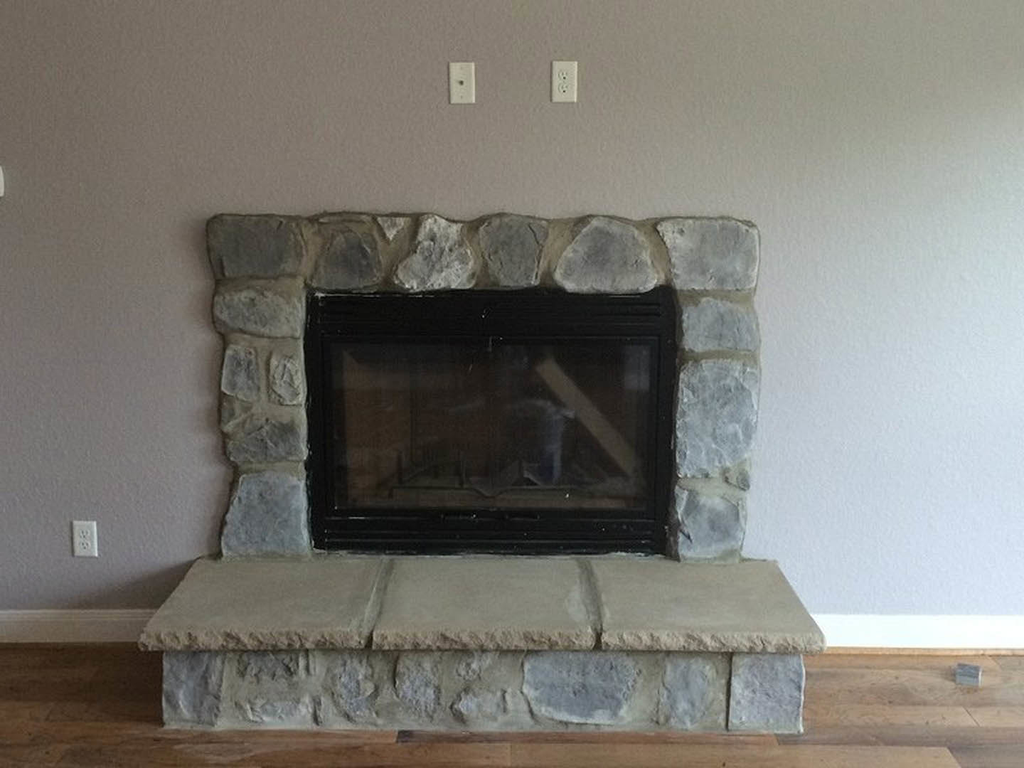 Stone fireplace with hearth and fire screen set against a textured stone wall, wood flooring, nearby window with glass pane, and white electrical outlet visible on the wall