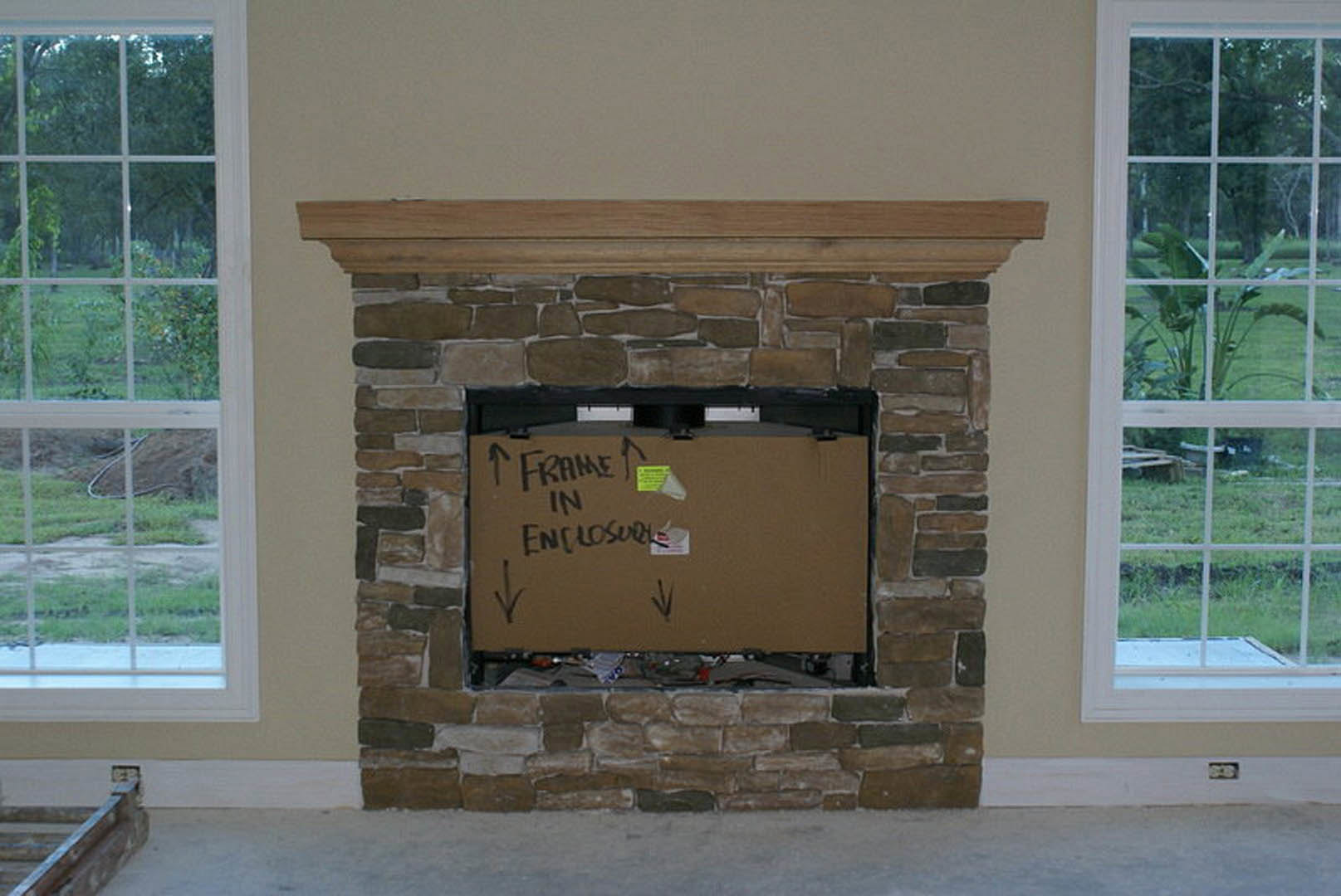 Stone fireplace with a cardboard box labeled in black text placed inside, wood flooring in foreground, large window showing green garden and plant outside