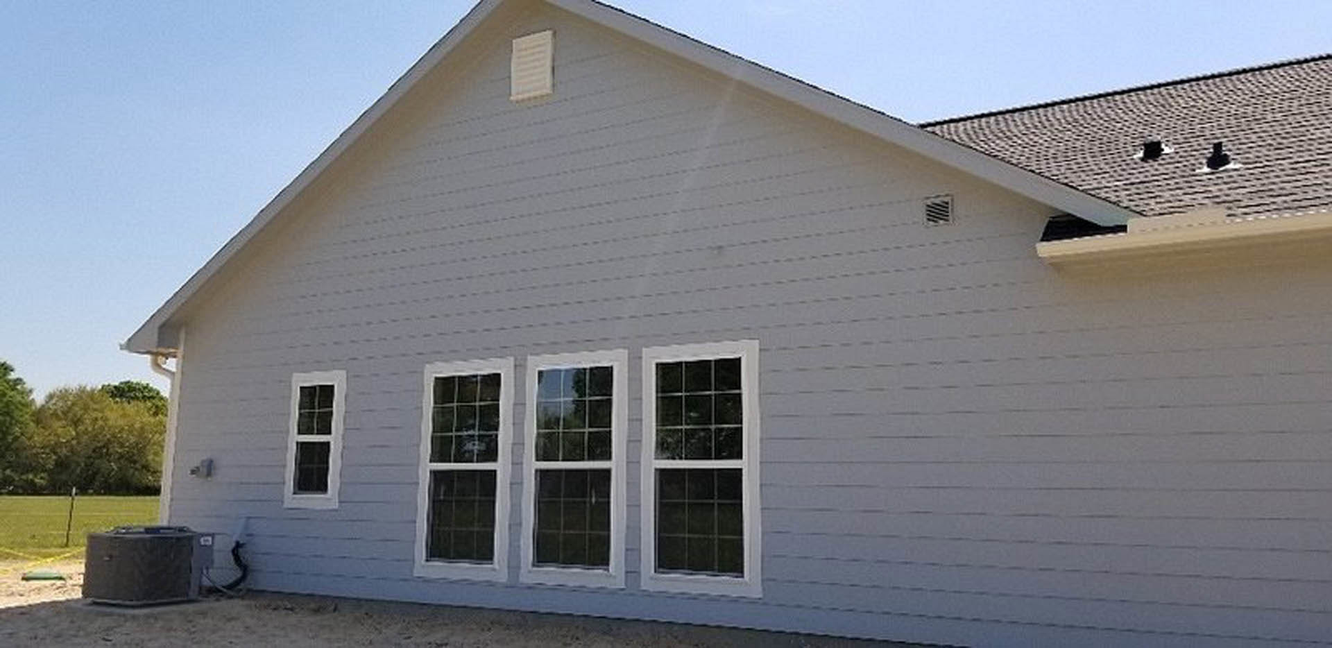 Modern cottage-style home with light-colored siding, dark roof, and minimal windows; entry door visible, surrounded by landscaped outdoor area under clear sky.