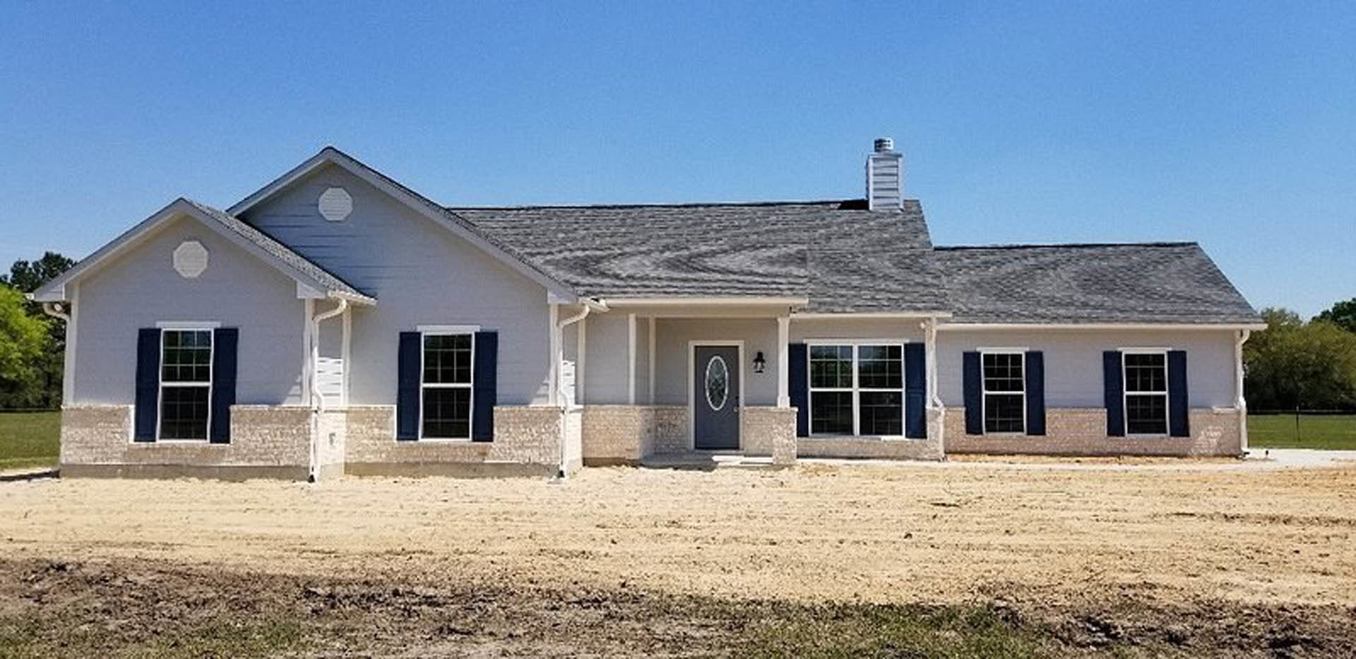 Two-story cottage with light siding, dark roof, multiple windows, and a bare dirt yard surrounded by sparse trees under a clear sky