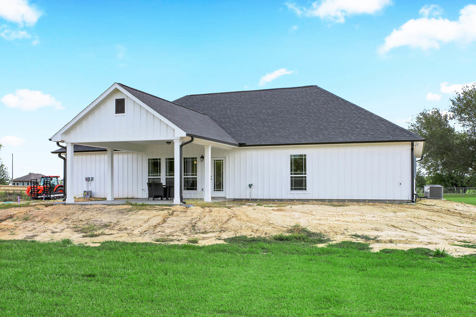 White house with black roof, white-framed windows, green lawn, dirt patch, red tractor with black metal rack, scattered trees under partly cloudy sky