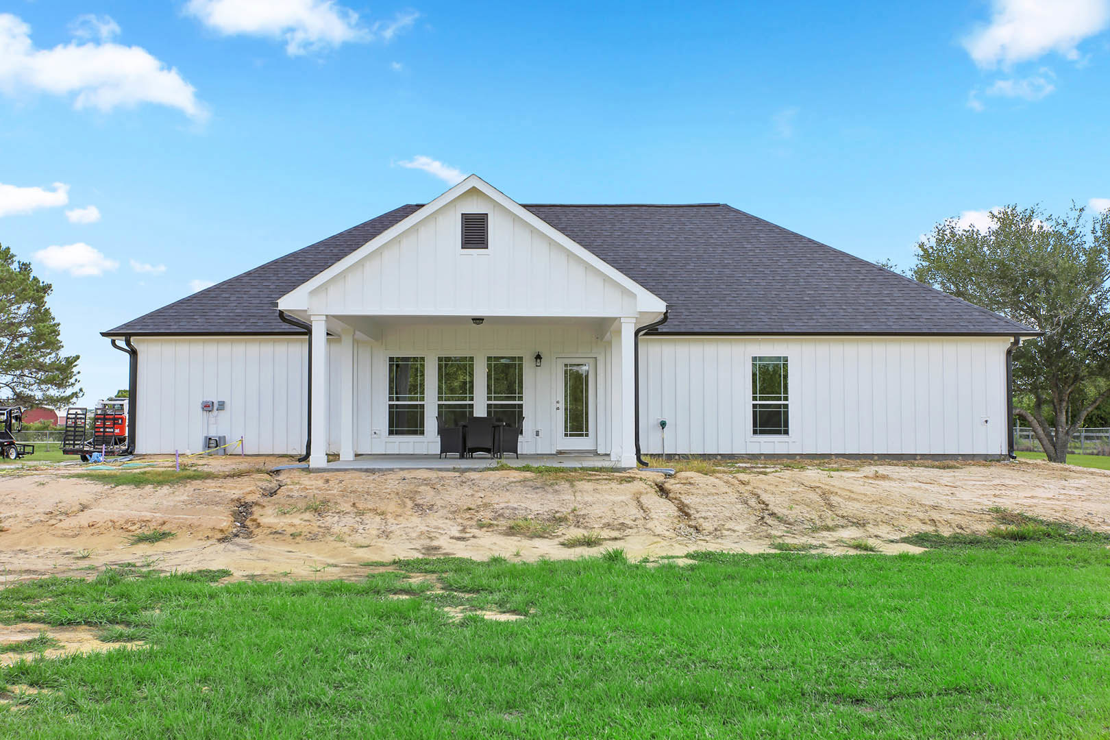 White two-story house with black roof, white-framed windows, and manicured green lawn; outdoor table and chairs set near a wooden fence with black ladder; trees and cloudy sky in