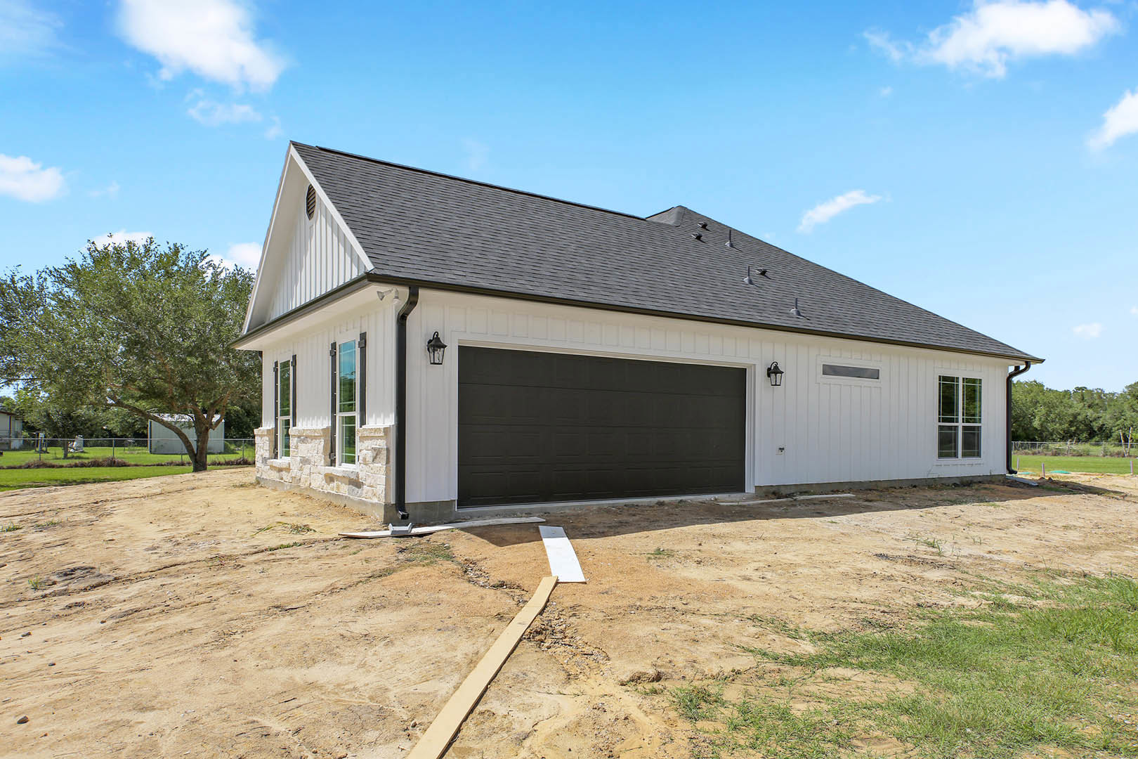 Partially built house with white siding, black-trimmed garage door, exposed wooden planks on dirt driveway, tree in front yard, cloudy sky overhead