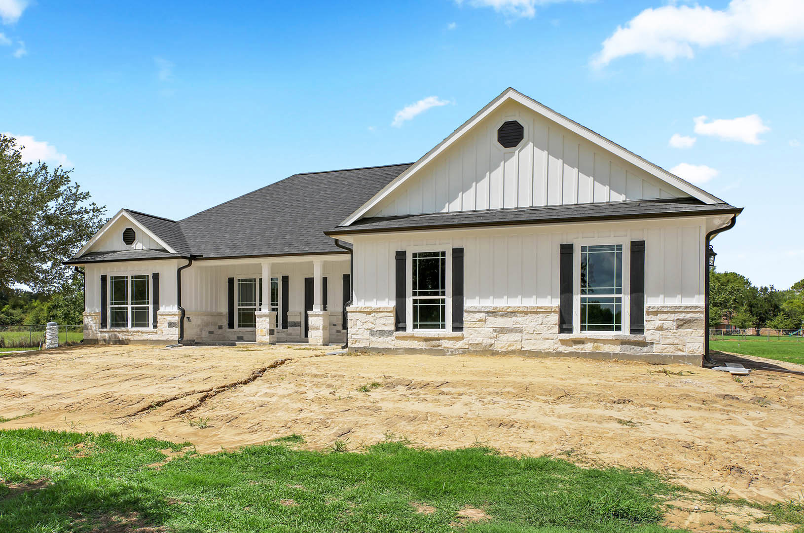 White house with black shutters and white-framed windows, black vent on exterior wall, dirt area in front, grass field beside building, trees and cloudy sky in background