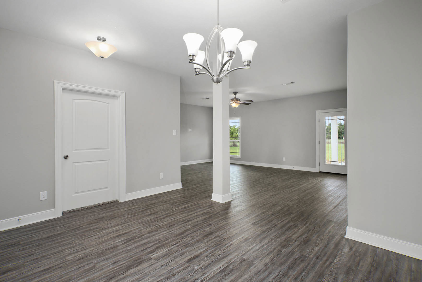 Wood flooring with white pillars, ornate chandelier hanging from plaster ceiling, white door with silver knob and vertical bars, neutral walls, ceiling fan visible in background