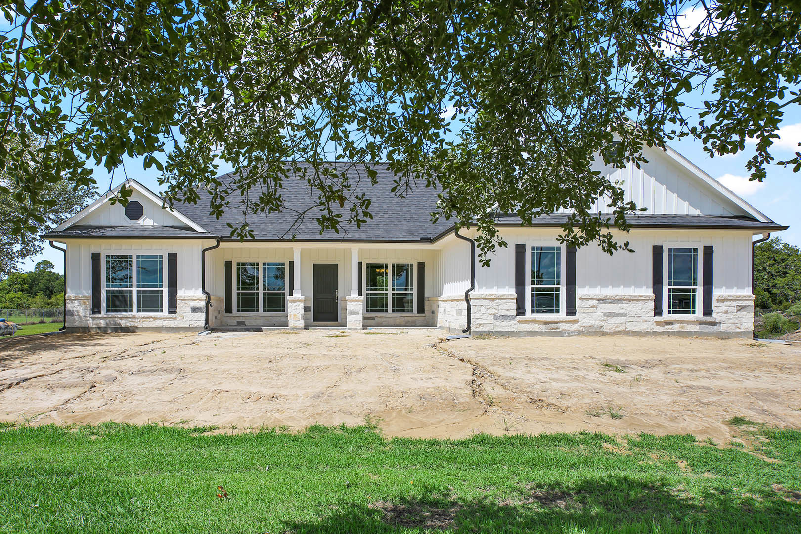 White house with black roof and windows, surrounded by trees, dirt road in foreground, white door with glass panes, grassy yard
