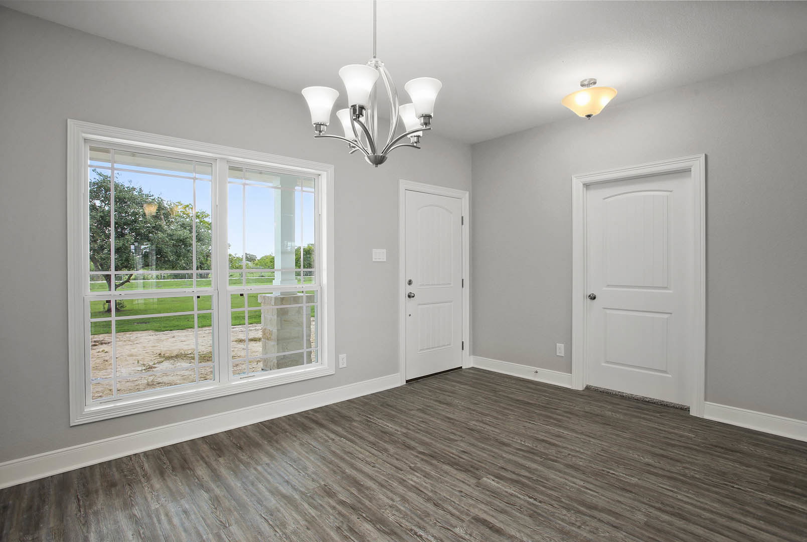 Chandelier hanging from ceiling in a room with wood flooring, white doors with silver handles, and large window overlooking yard