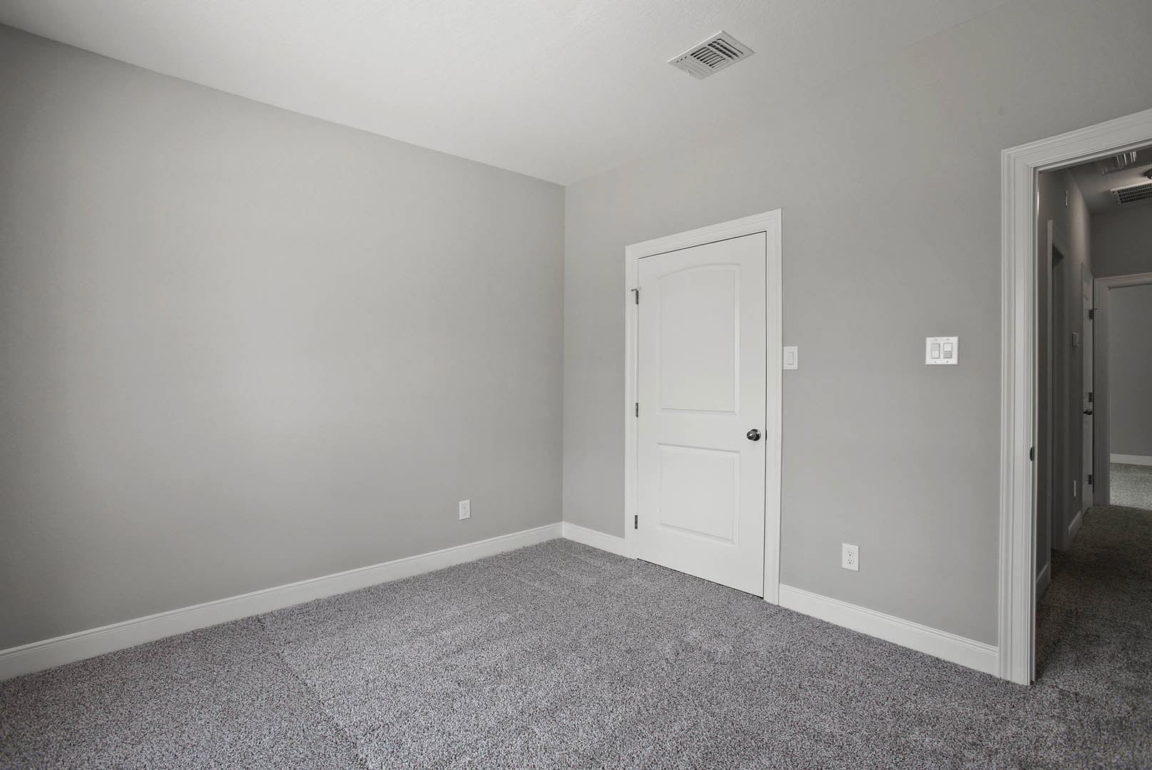 White paneled door with silver knob, beige carpet flooring, white walls with light switch, ceiling vent visible