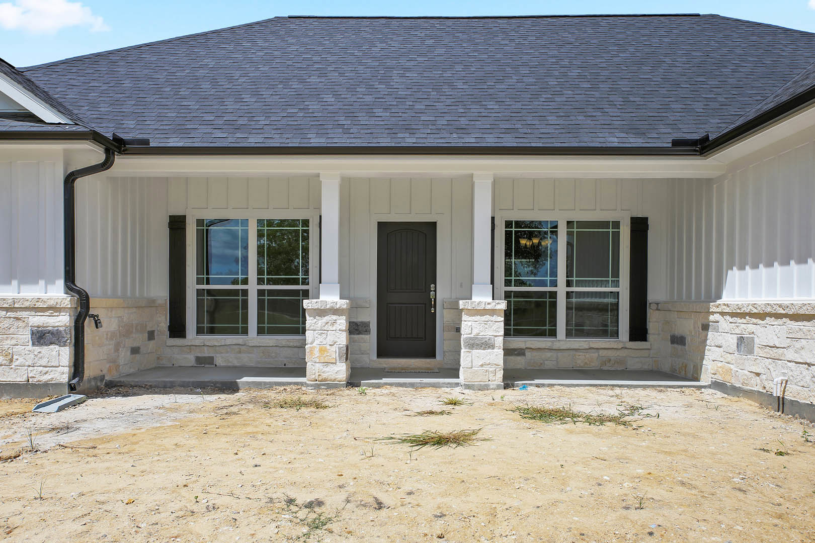 Black front door framed by stacked stone pillars, light siding, double windows, and shingled roof above entryway