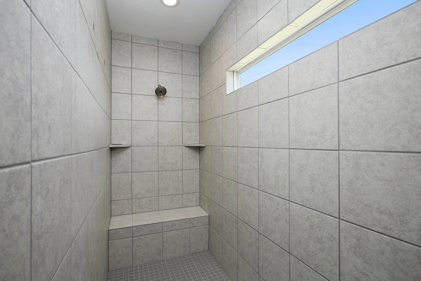 Walk-in shower featuring a large window, grey tile flooring with white grout, marble wall tiles, and modern ceiling-mounted shower head