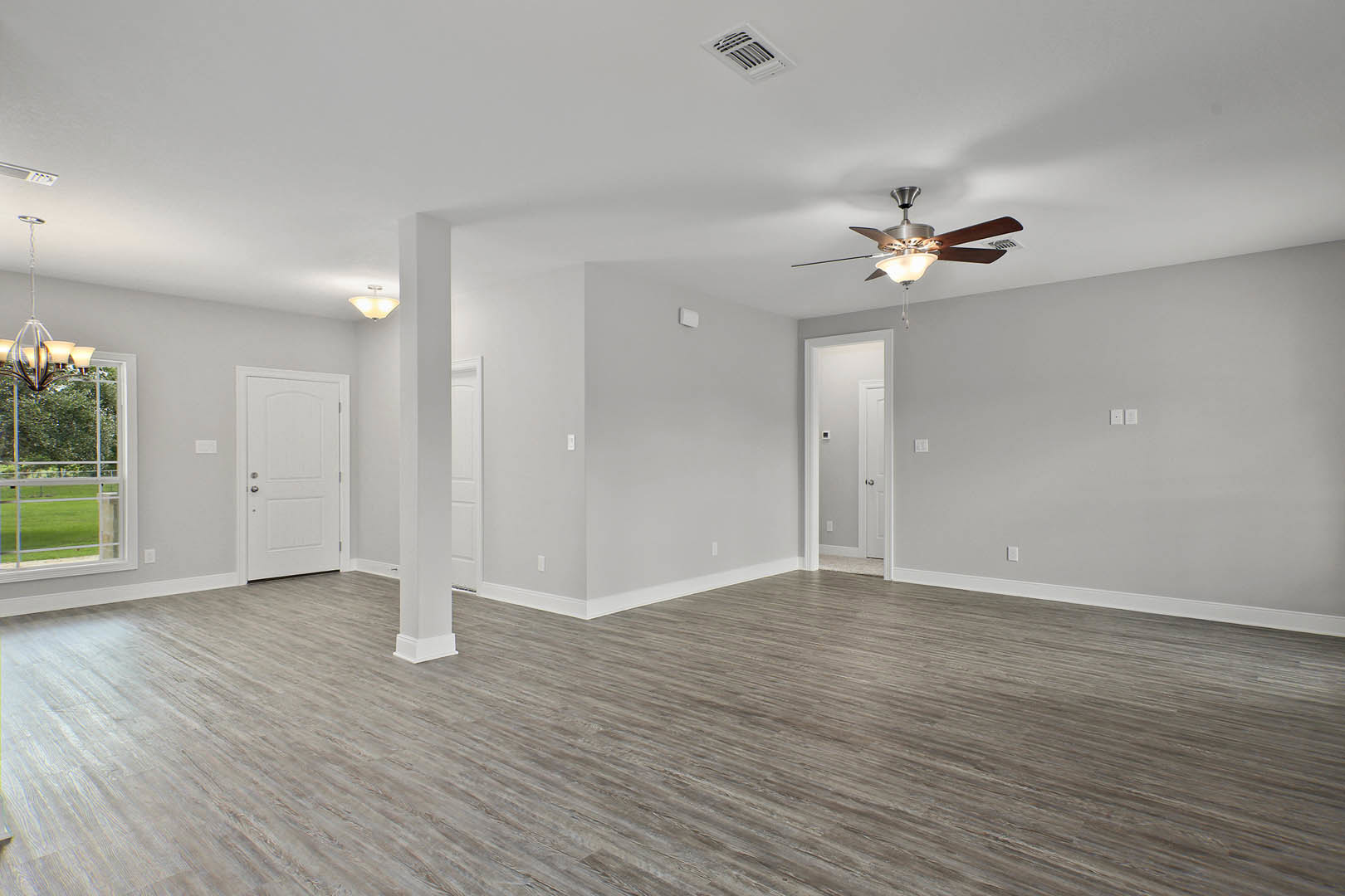 Ceiling fan with light fixture, wood flooring, white walls, white door with silver handle and black square accent, window overlooking green lawn